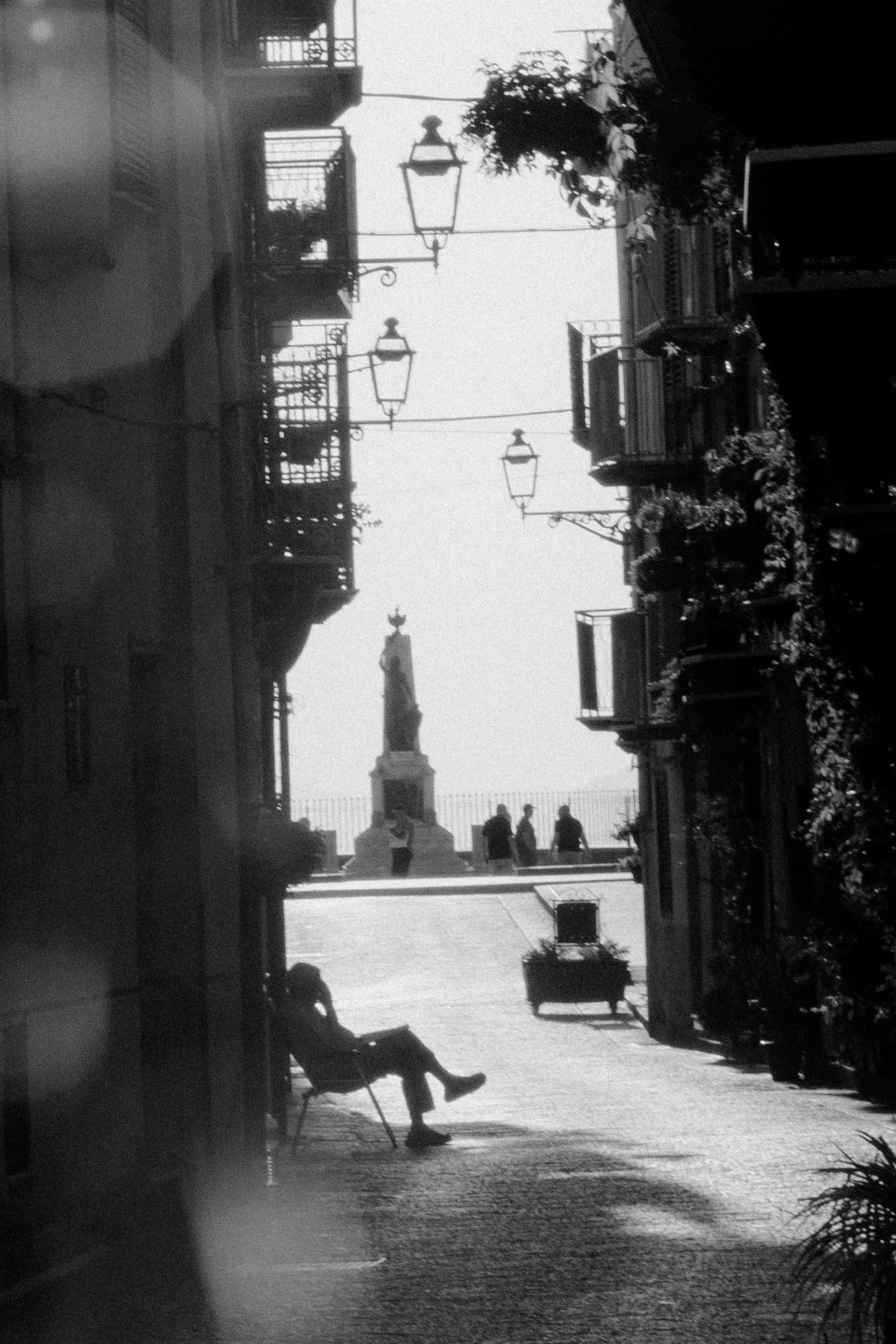 A black and white street scene in Italy with vintage lanterns and silhouetted figures against bright sunlit buildings.