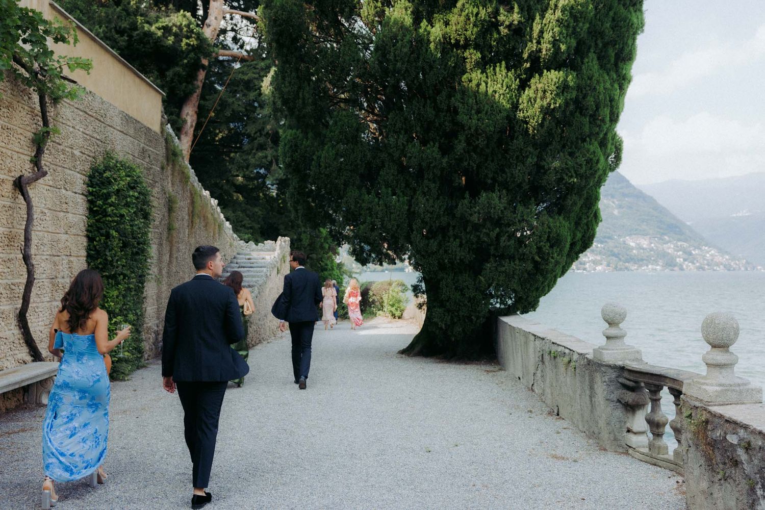 People walking along a scenic stone pathway with trees and mountain views in the background.