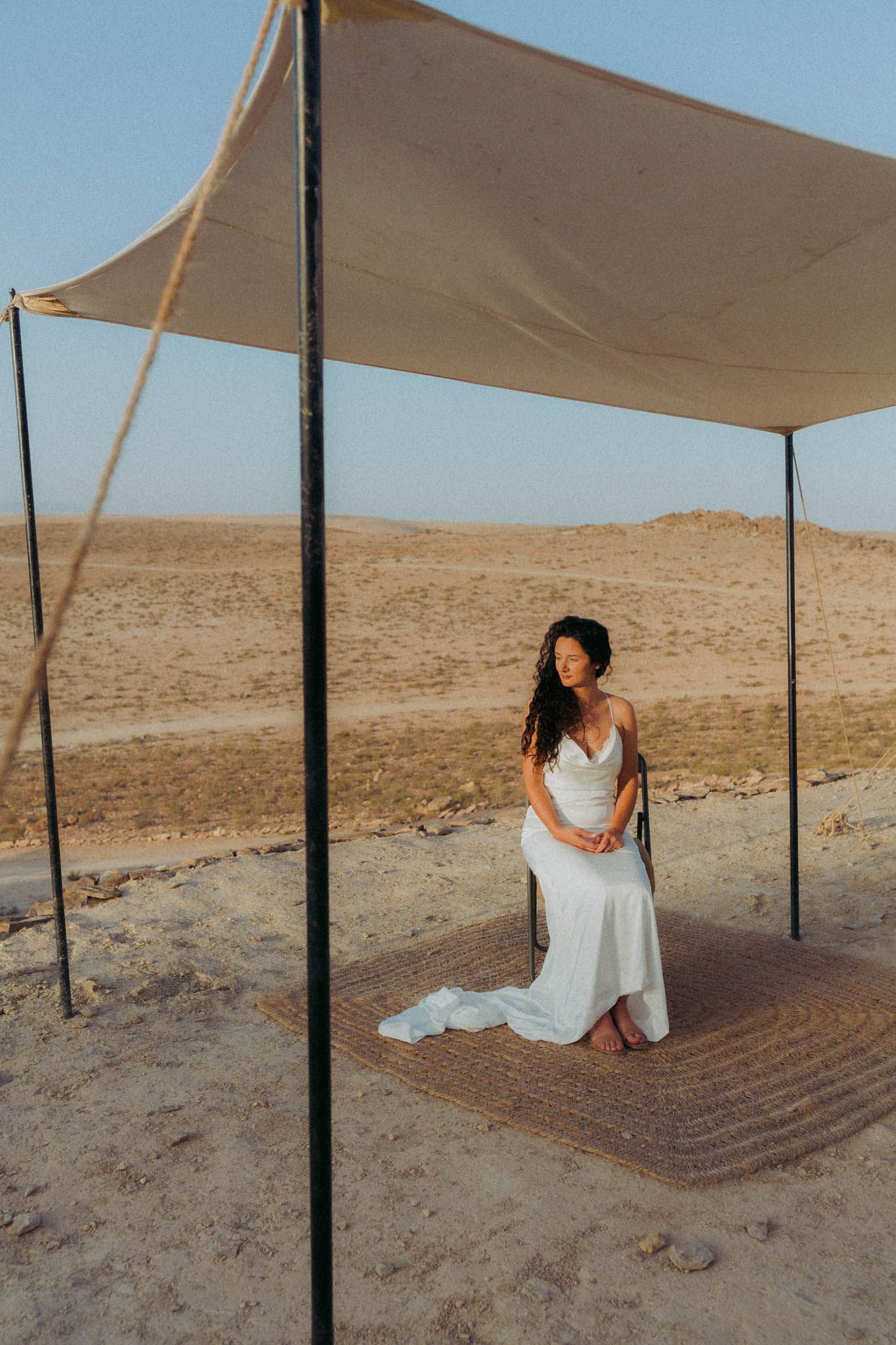 A person in a white dress poses under tan canopy shades in a desert landscape during a photoshoot sequence.