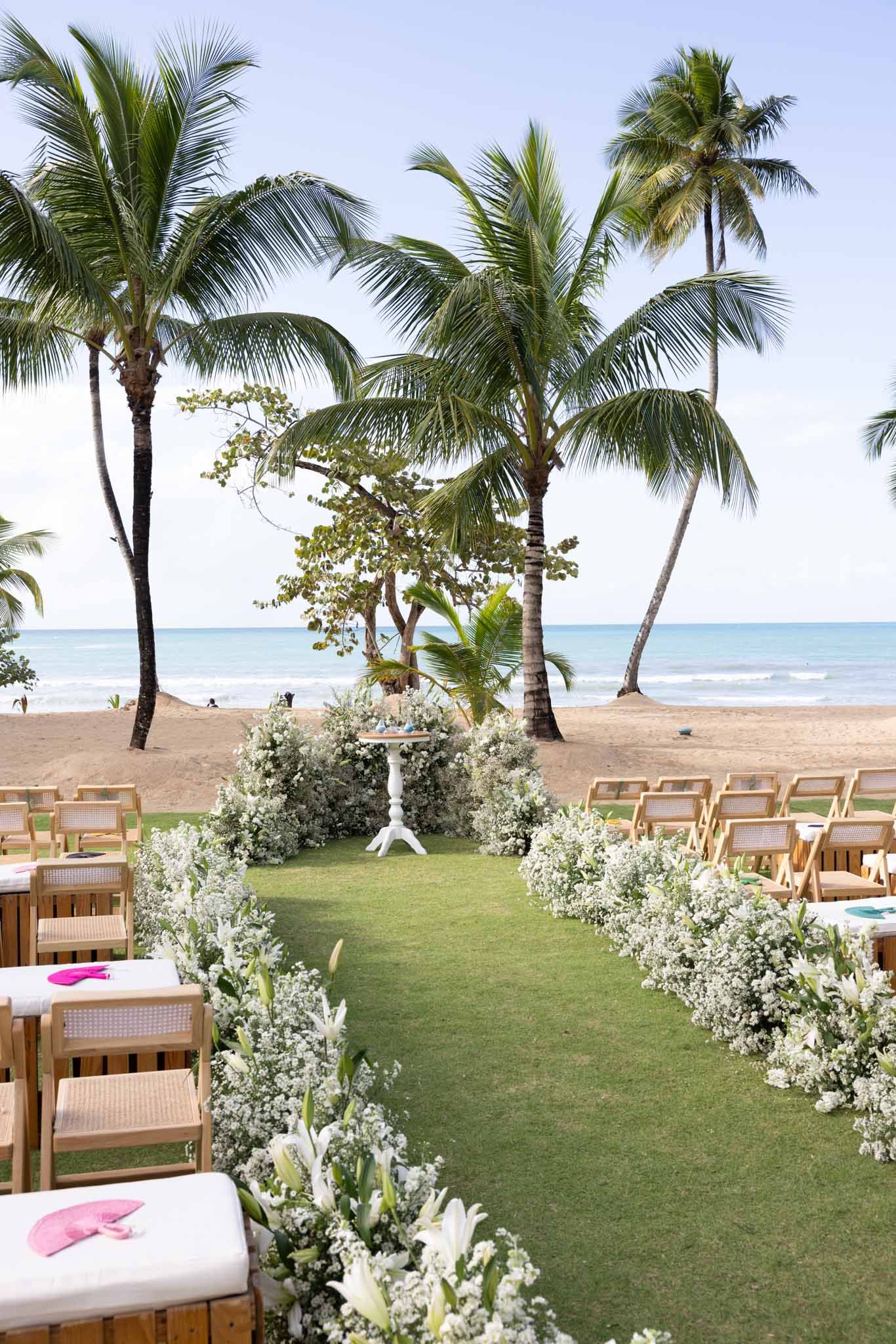 Tropical beach wedding ceremony setup with wooden chairs, white floral aisle decorations and palm trees overlooking the ocean.