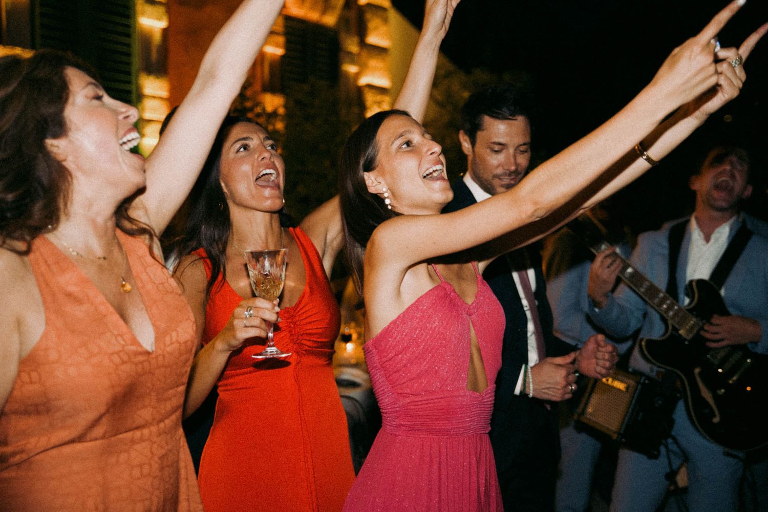 Enthusiastic party guests in colorful dresses dance with raised arms while celebrating at a nighttime event.