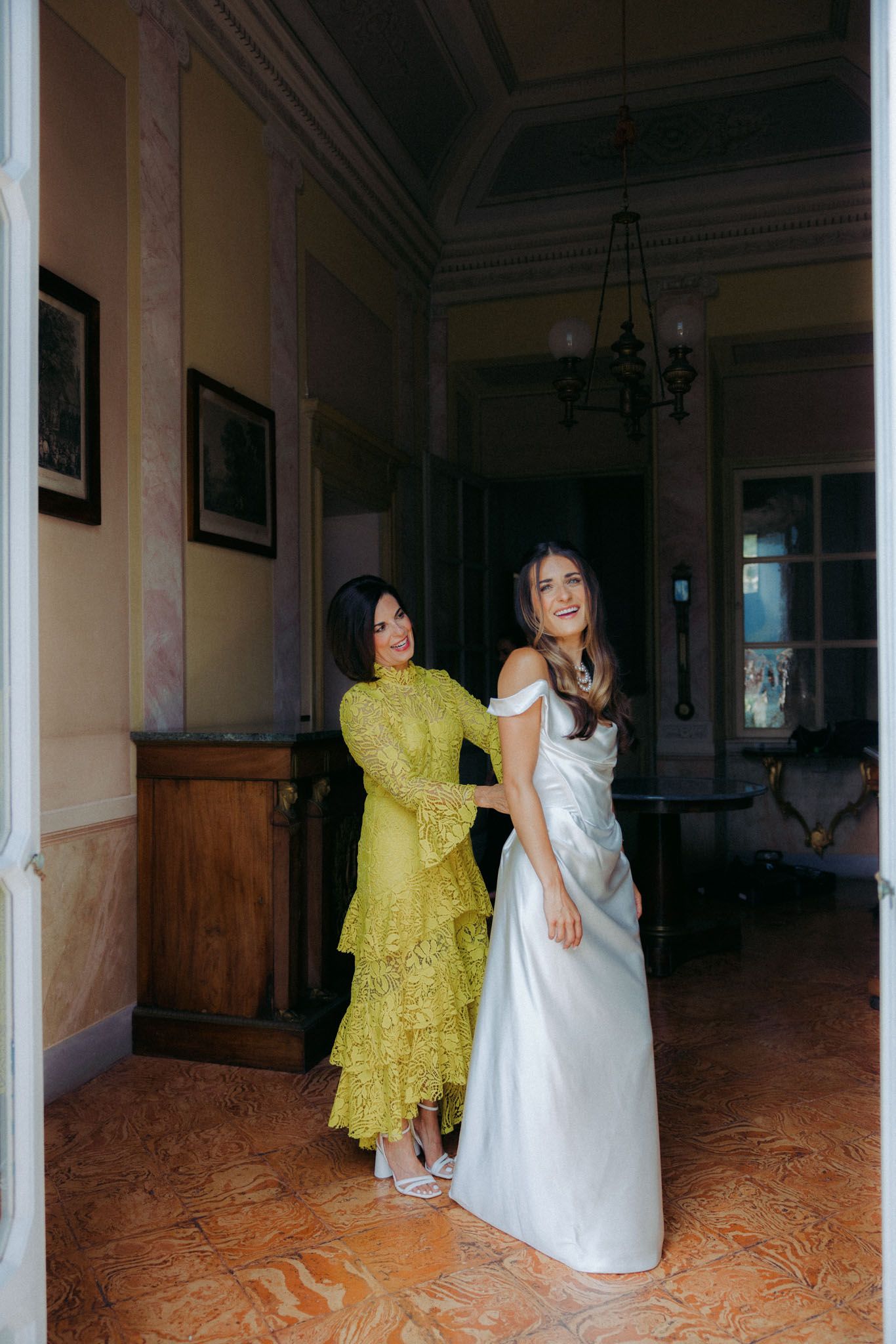 Two women pose in elegant dresses - one in yellow lace and one in white satin - inside a grand hallway with ornate details.