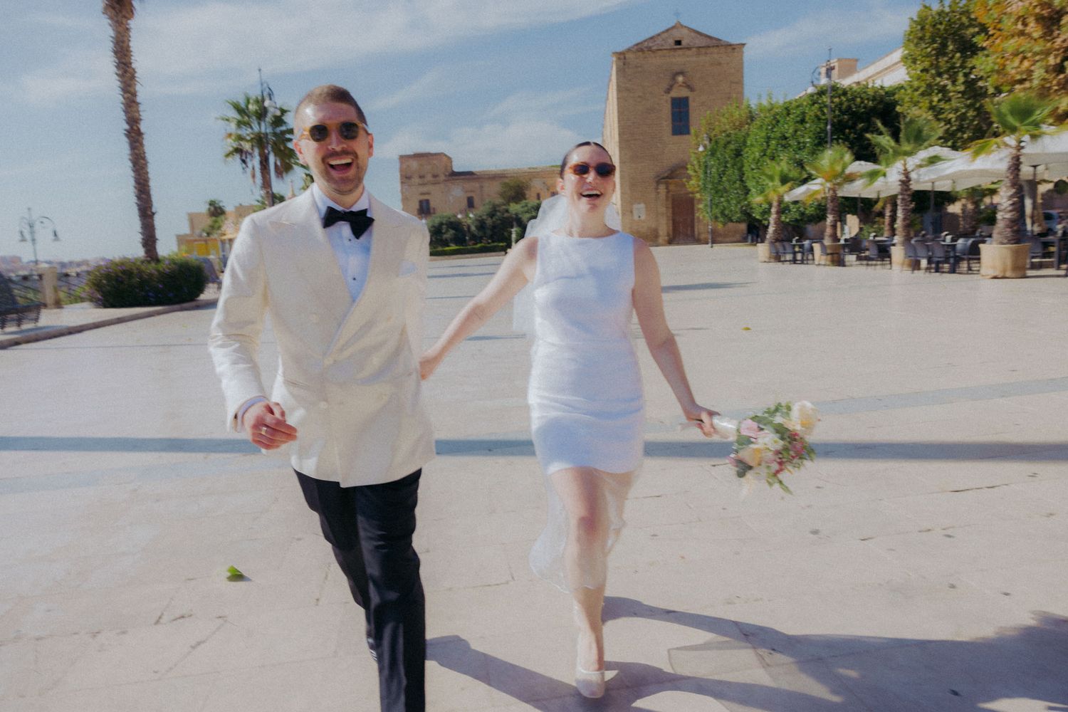 Couple walking hand in hand on a sunny day in an outdoor plaza with stone buildings in the background.