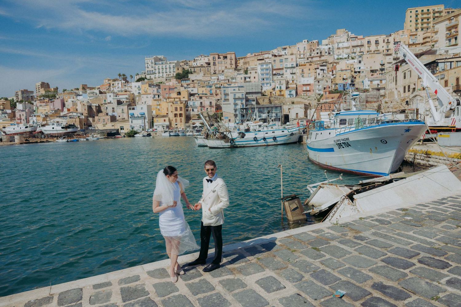 Mediterranean waterfront promenade with historic buildings and boats docked in the harbor on a sunny day.