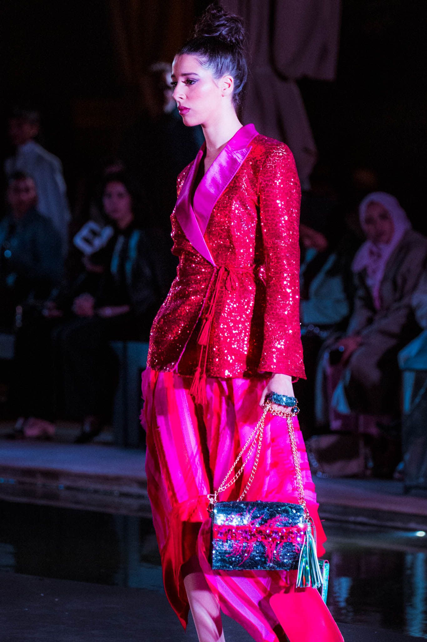 Model walks runway in bright pink sequined dress with glittering handbag during fashion show.