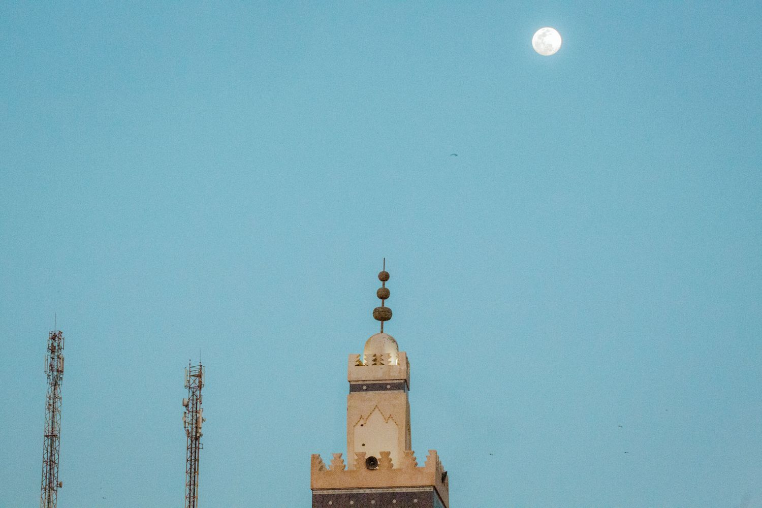 A full moon rises over a desert landscape with silhouetted buildings and towers against a blue twilight sky.