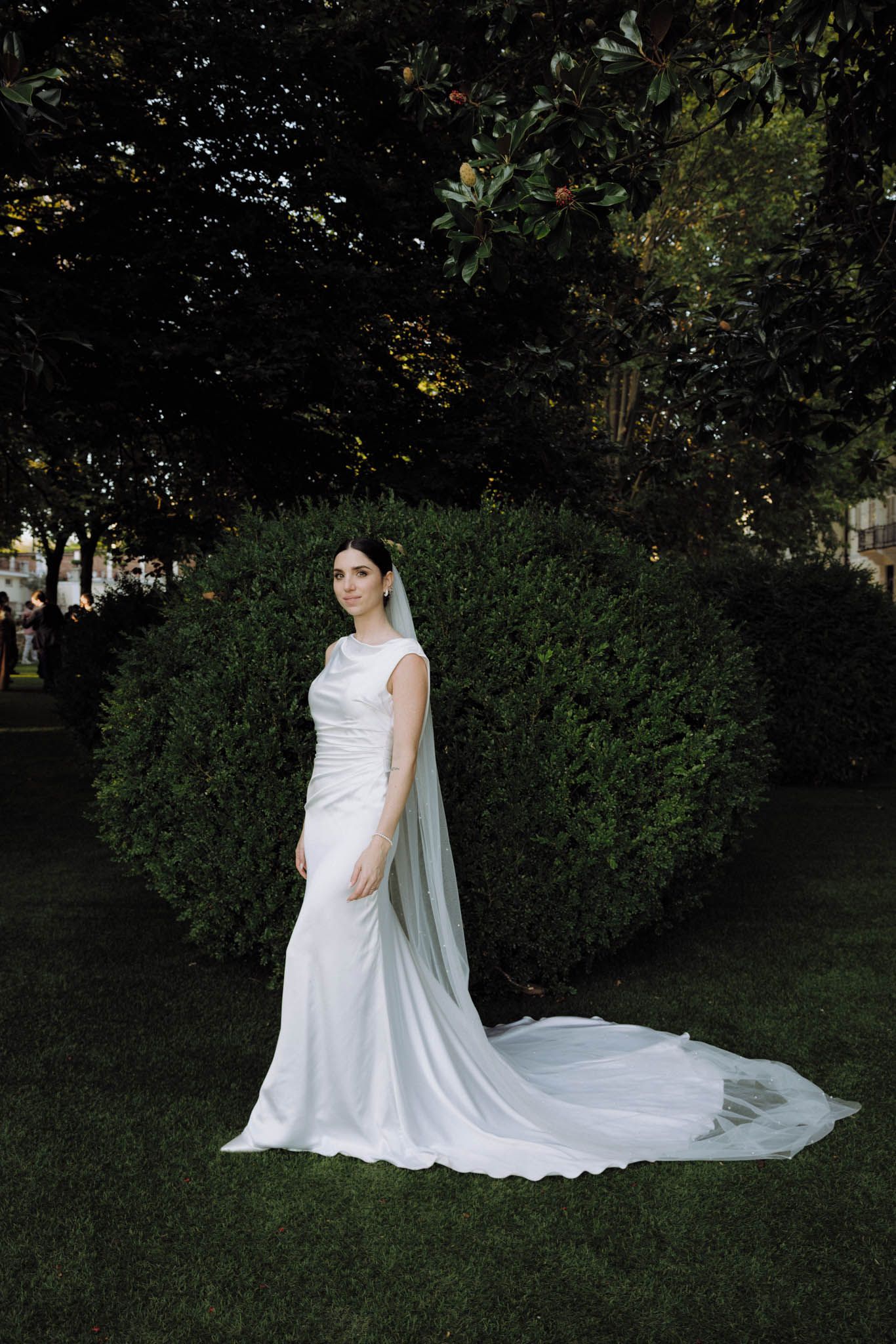 A flowing white wedding dress with a long cathedral veil creates an elegant silhouette against a garden backdrop.