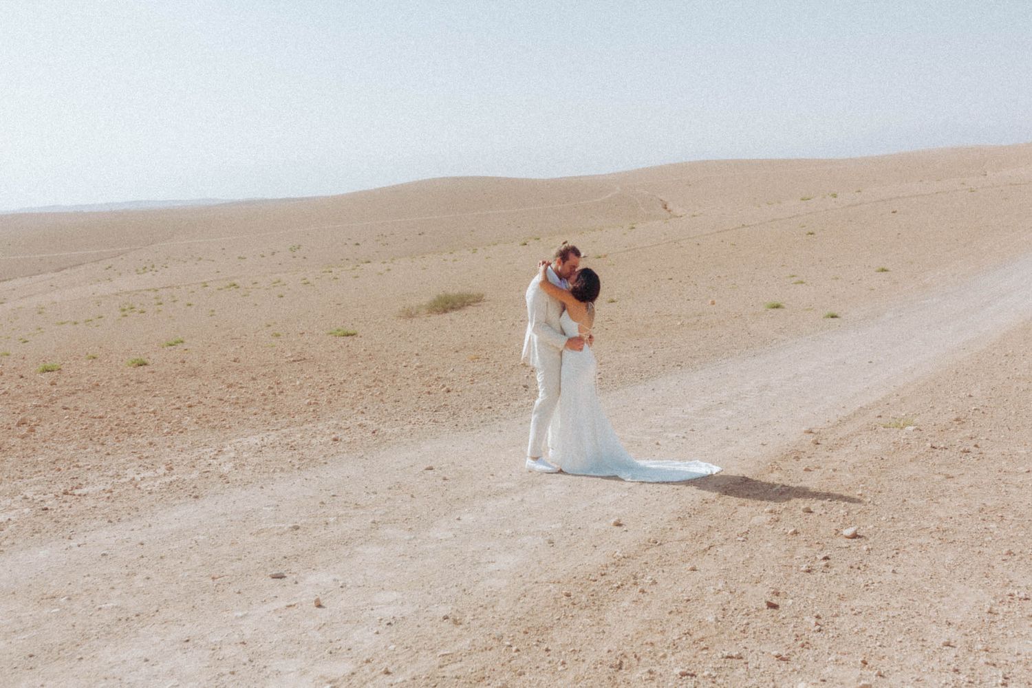 A couple in wedding attire embraces while standing on a winding desert road beneath a hazy sky.