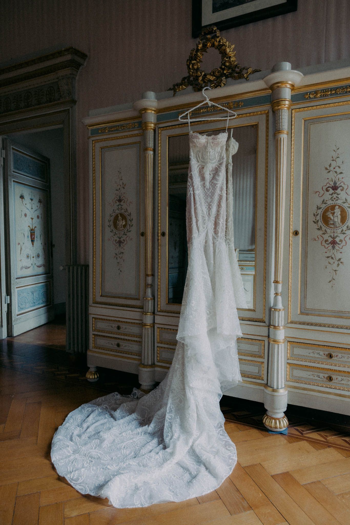 White lace wedding dress displayed on a hanger in an elegant room with ornate gold trim and floral wall details.