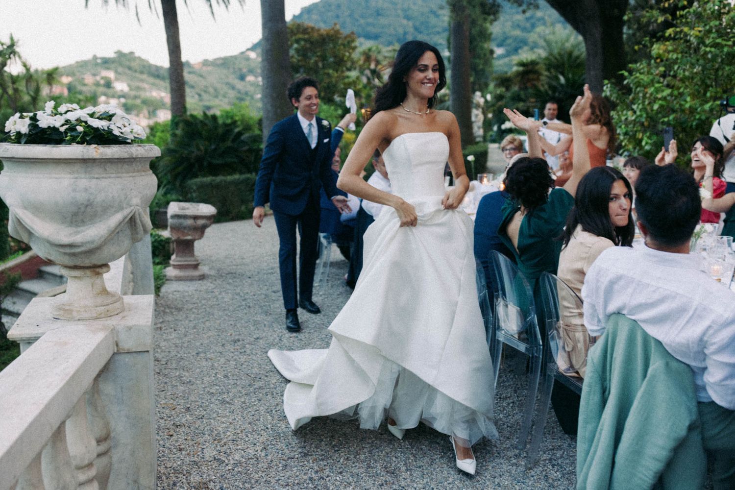 Wedding guests dance and celebrate at an elegant outdoor reception with stone architecture and lush greenery.