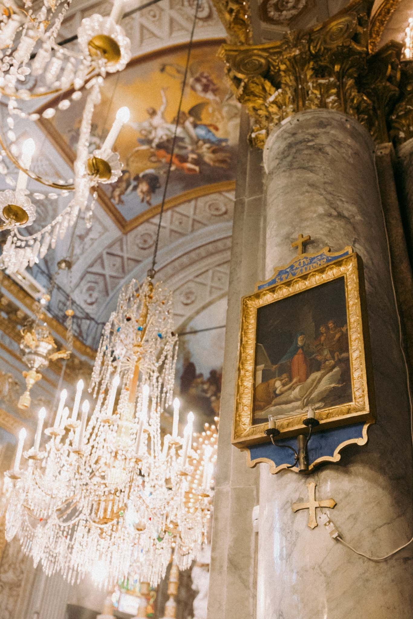 Ornate crystal chandeliers hang from an elegant baroque ceiling with gilded frames and decorative moldings in a palace interior.
