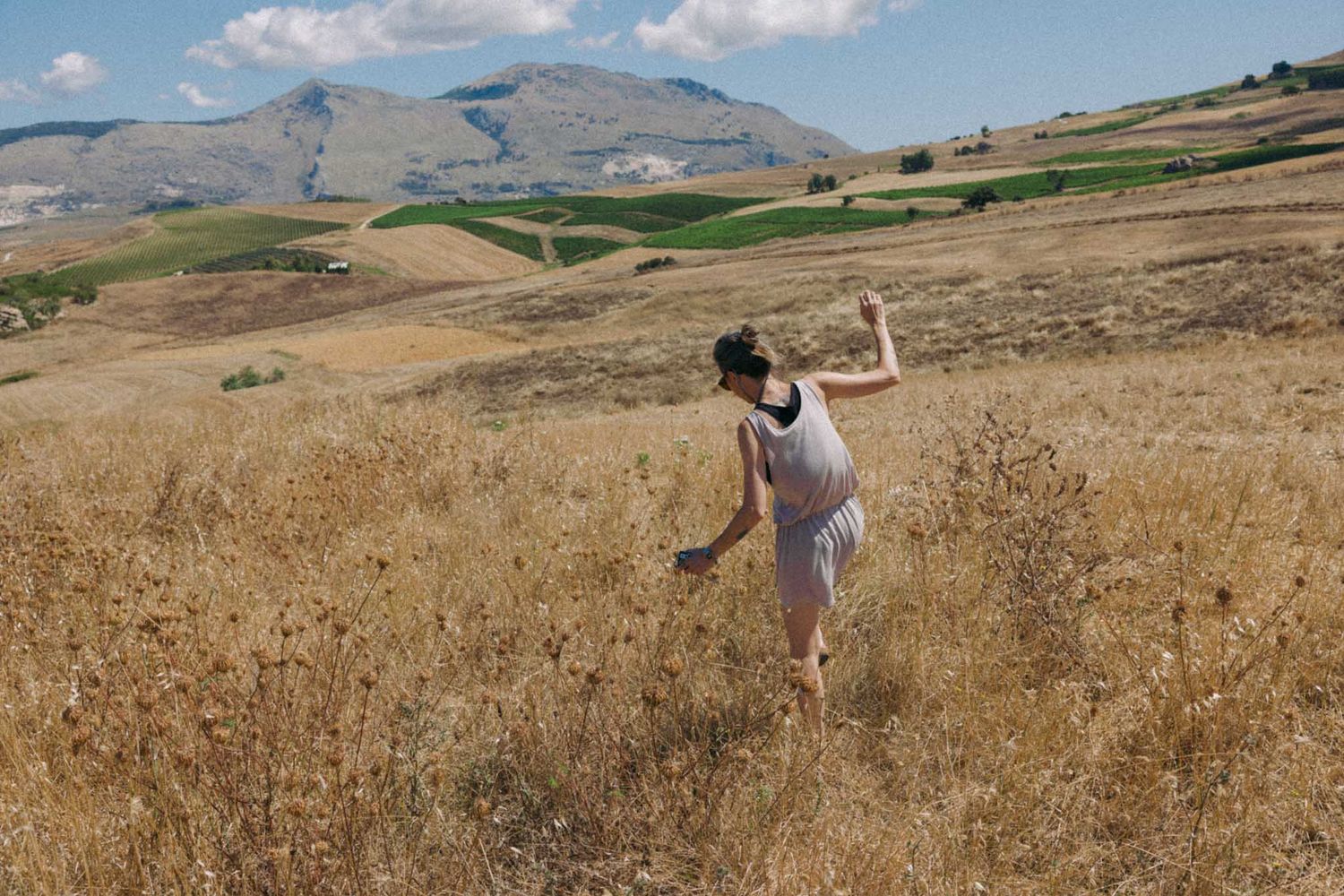 Person walking through a dry golden field with rolling hills and blue sky in the background.