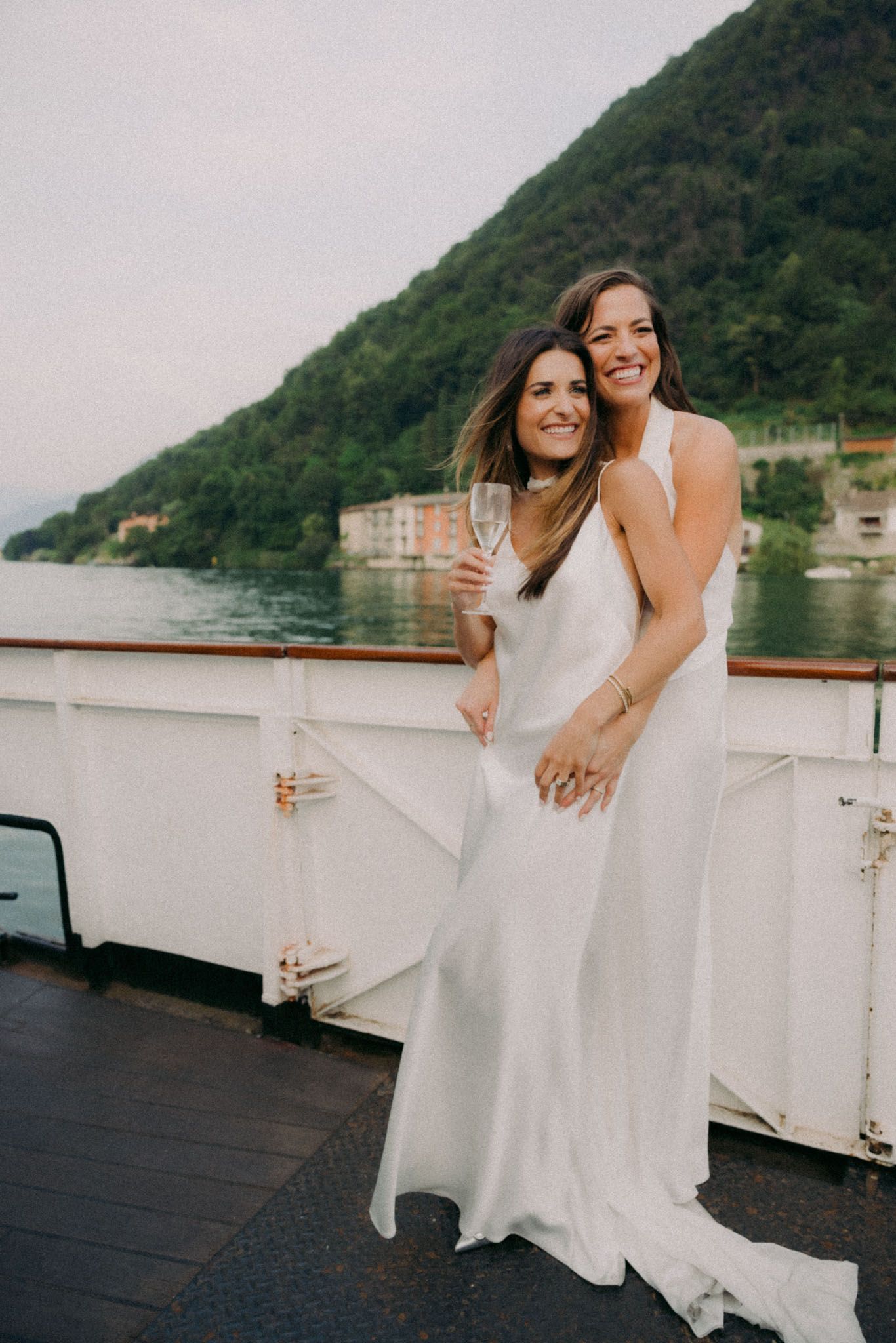 Two people in white dresses share a joyful moment on a boat deck with scenic mountains and waterfront buildings behind them.