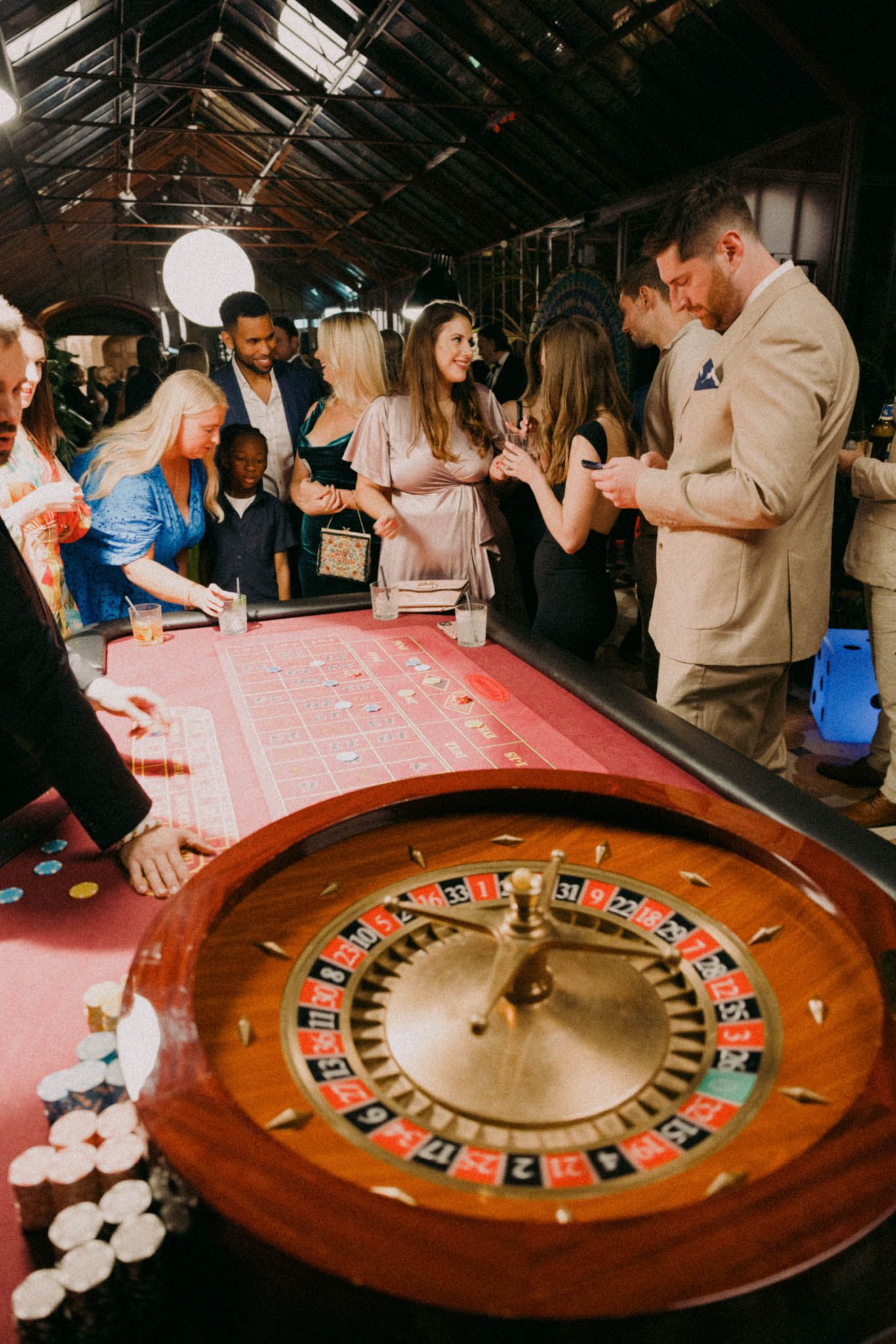 People gather around a casino roulette table with red felt and wooden trim during a social event.