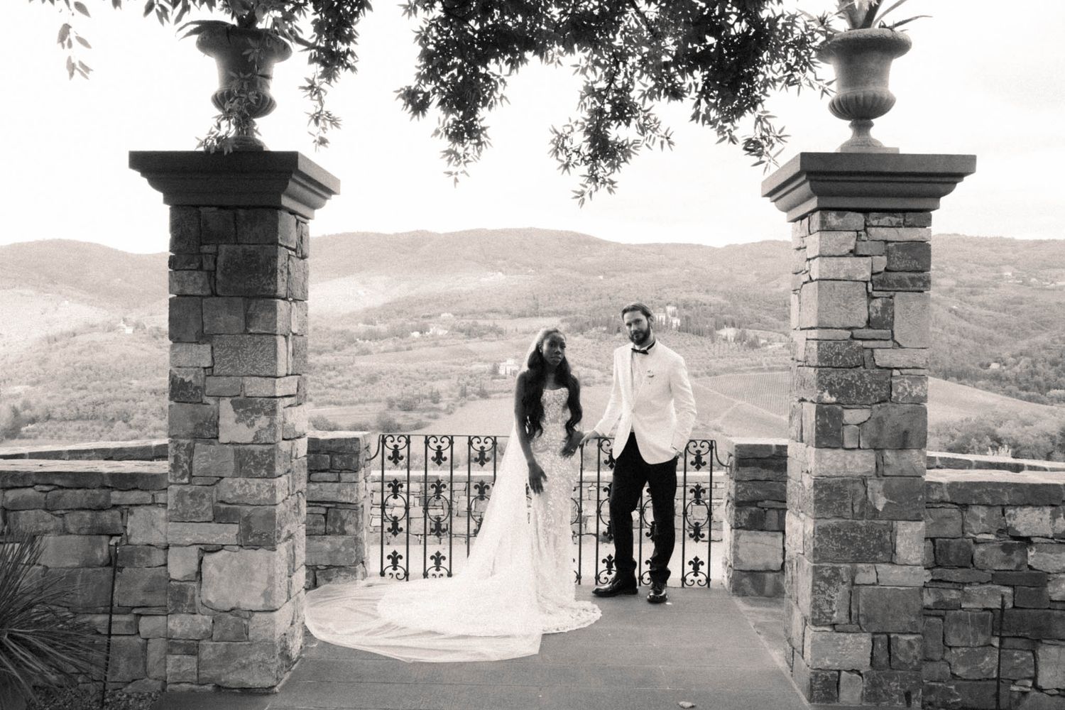A black and white panoramic view of stone pillars and gates at a historic estate entrance with mountains in the background.