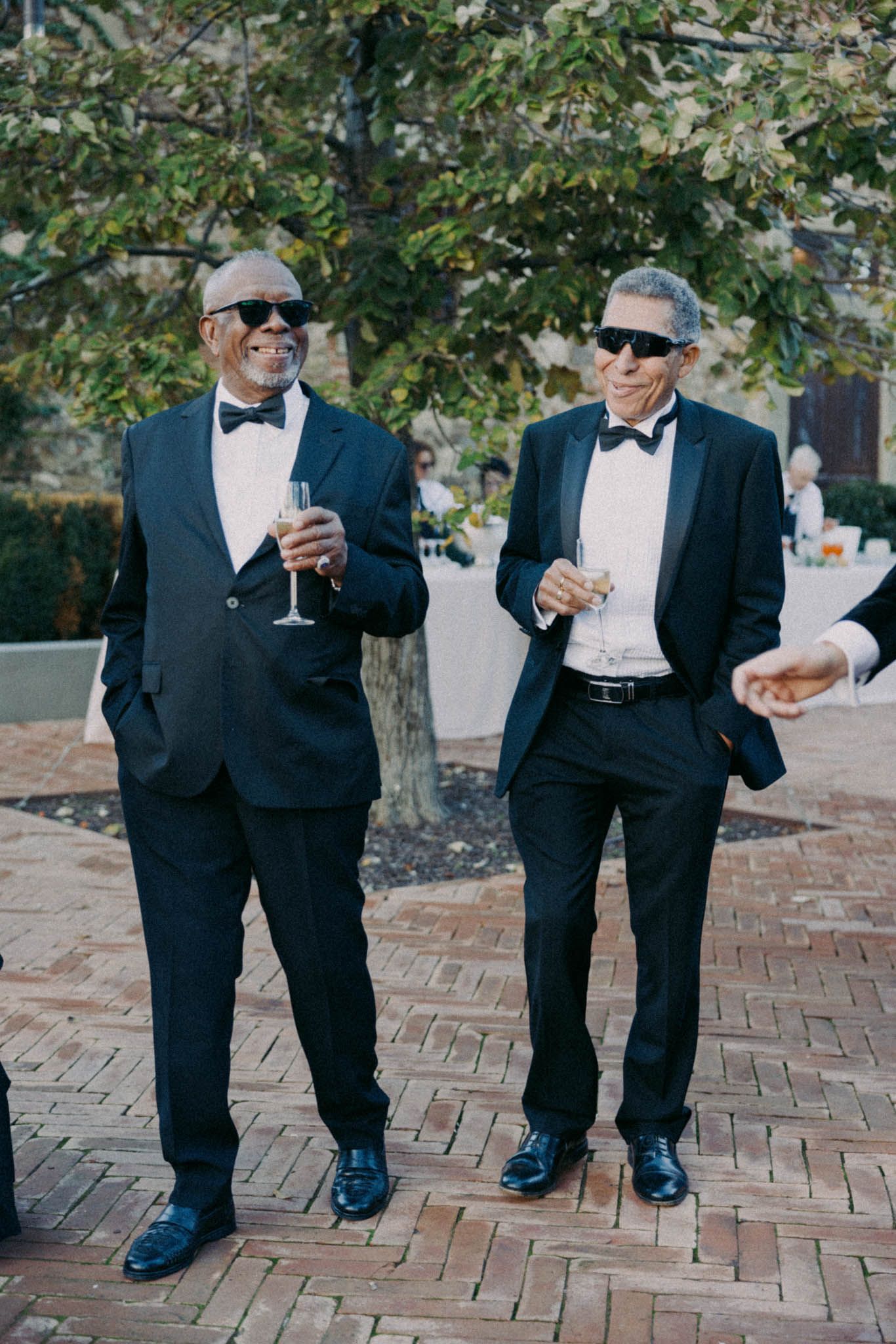 Two people in formal black tuxedos and bow ties share a lighthearted moment on a brick patio.
