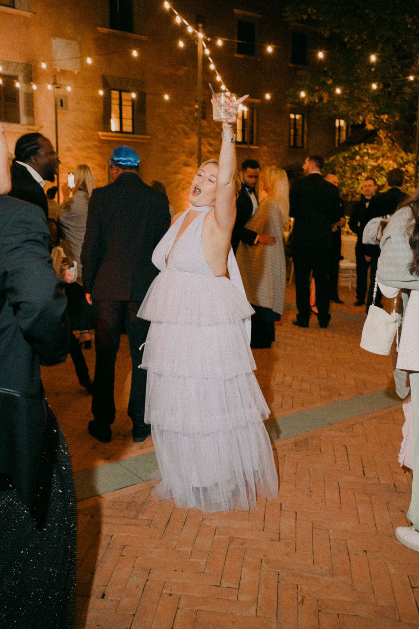 Wedding guests mingle on a brick patio at night under warm string lights while a bride in white dress stands nearby.