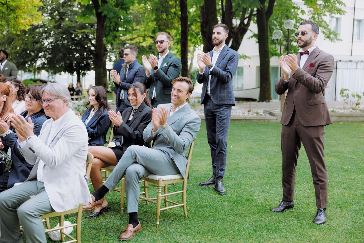 A group of people sit in chairs arranged in a circle outdoors on a grassy lawn during a social gathering.