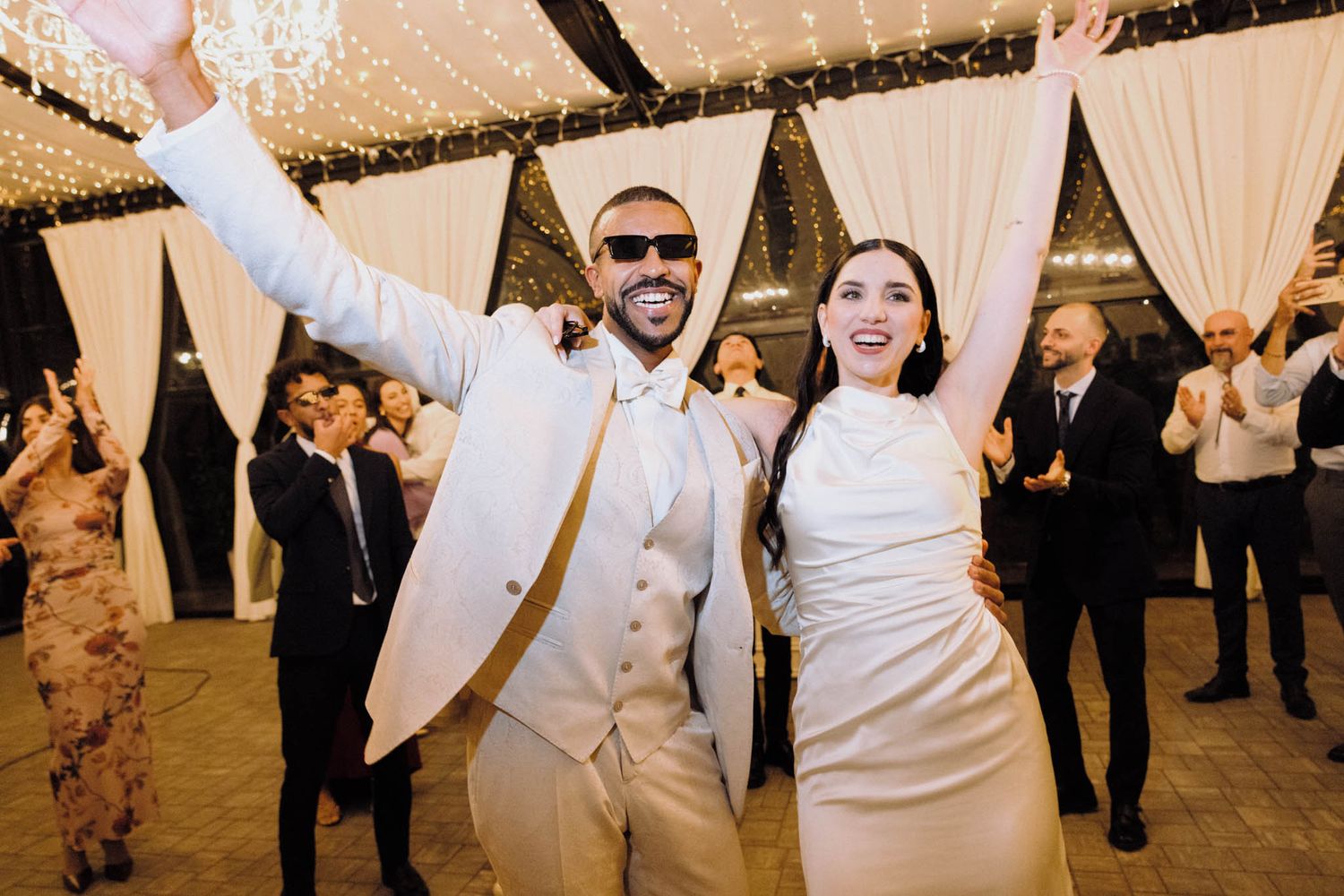 Wedding reception guests dance and celebrate under white draped tent decorations at an evening party.