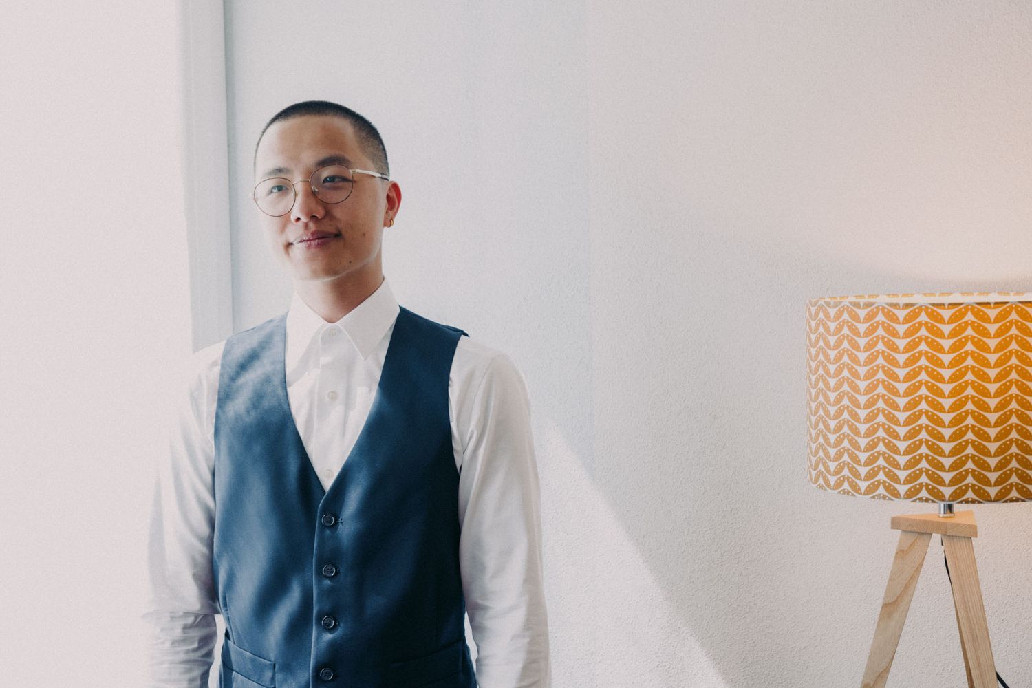 A person in a navy blue vest and white shirt stands against a light colored wall.