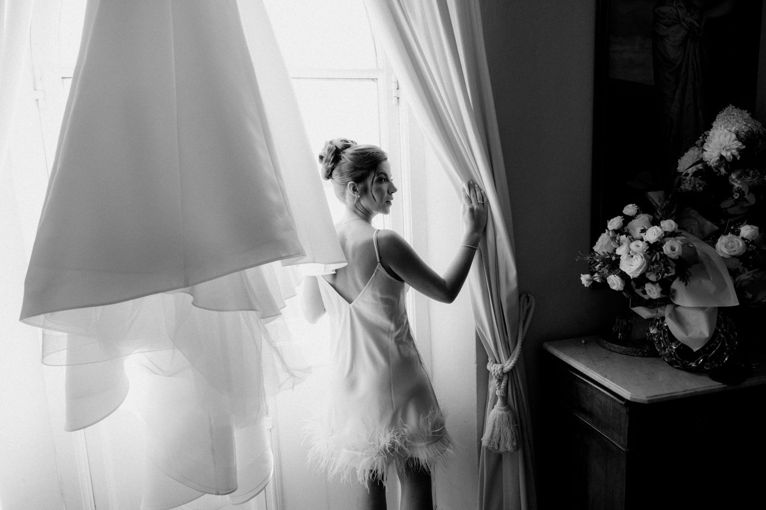 A row of white wedding dresses hanging by windows in a bridal boutique with natural light streaming through.