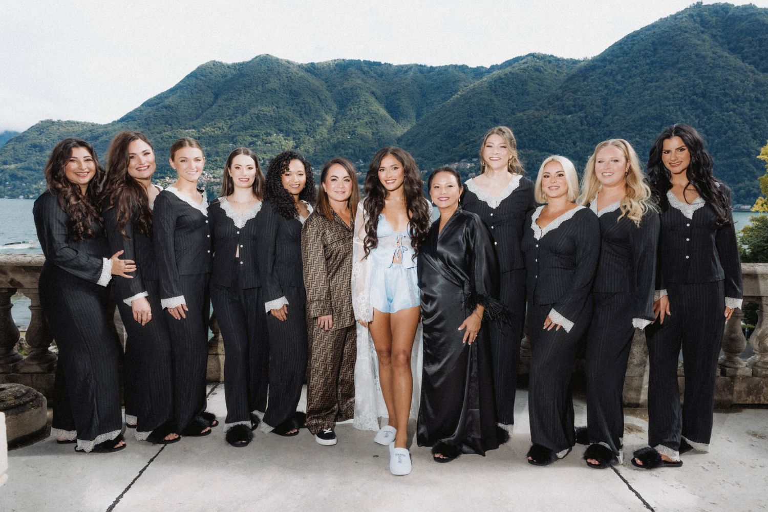 A group of people wearing black formal attire and one in white stand together on a terrace overlooking mountains and water.