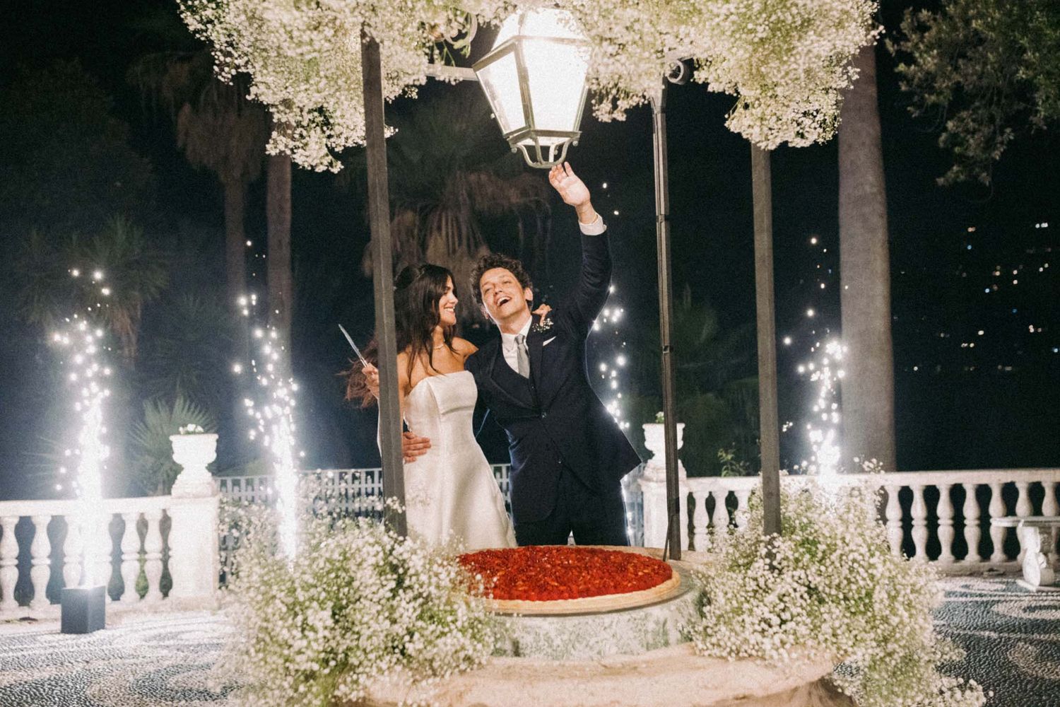 A couple shares a romantic dance moment under twinkling lights and blossoming trees in an outdoor evening setting.