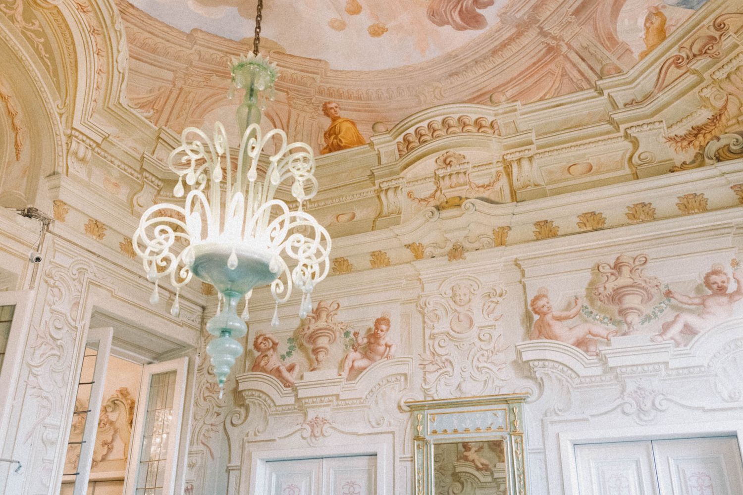 Ornate baroque palace interior with white and gold decorative moldings, frescoed ceilings, and crystal chandeliers.