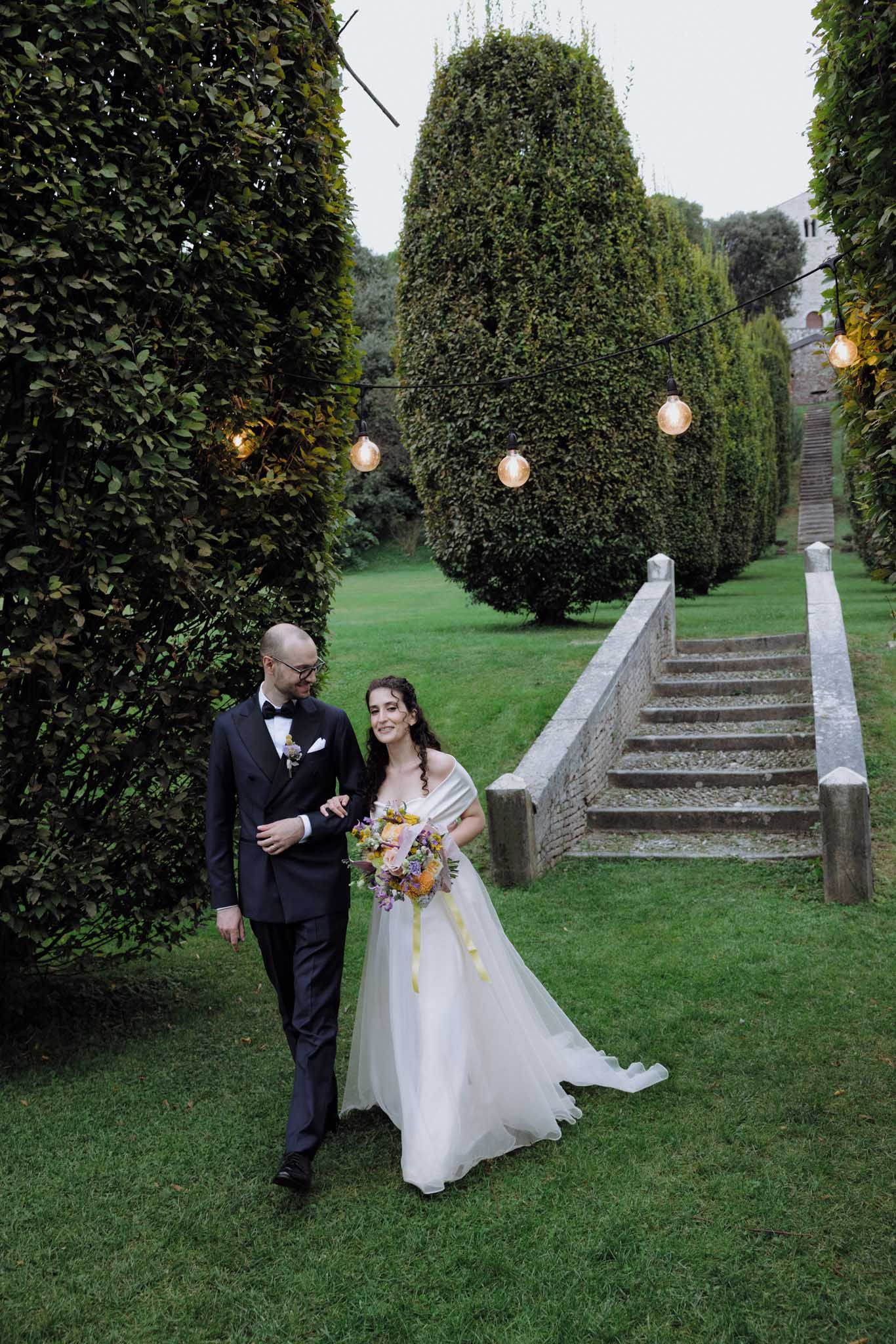 A couple in formal wedding attire walks through a garden path lined with topiaries and string lights at dusk.