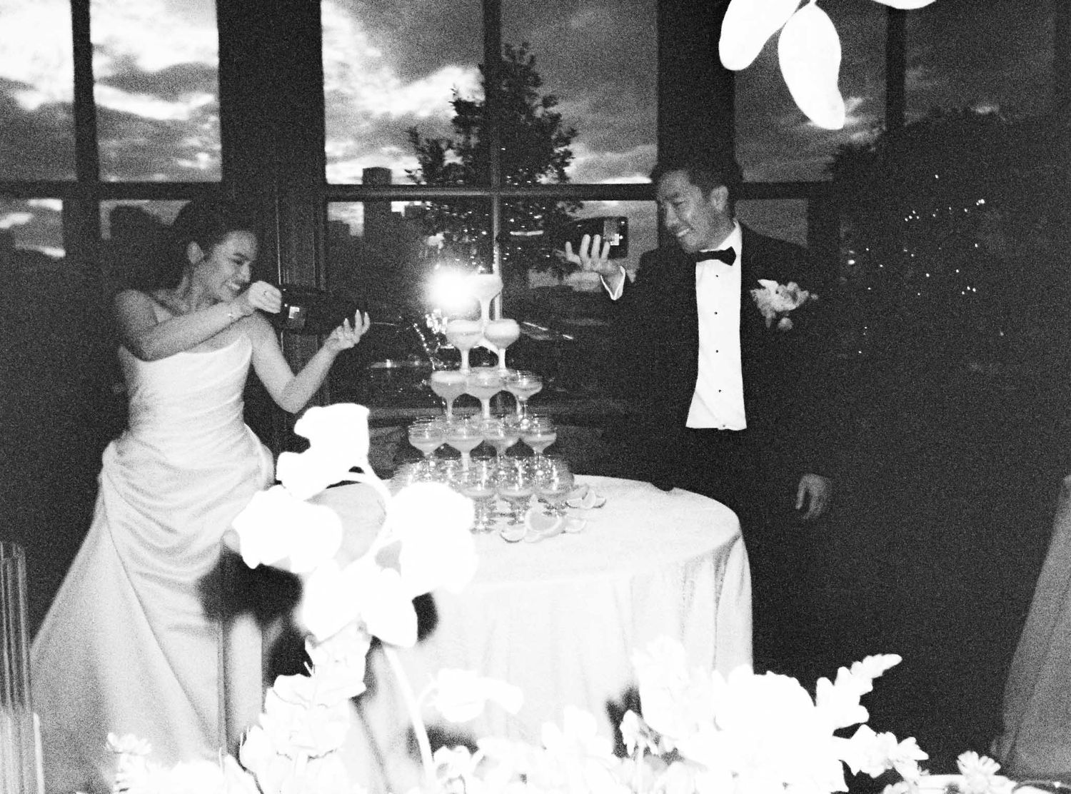 A couple pours champagne into a tower of glasses at their wedding reception in a black and white photograph.