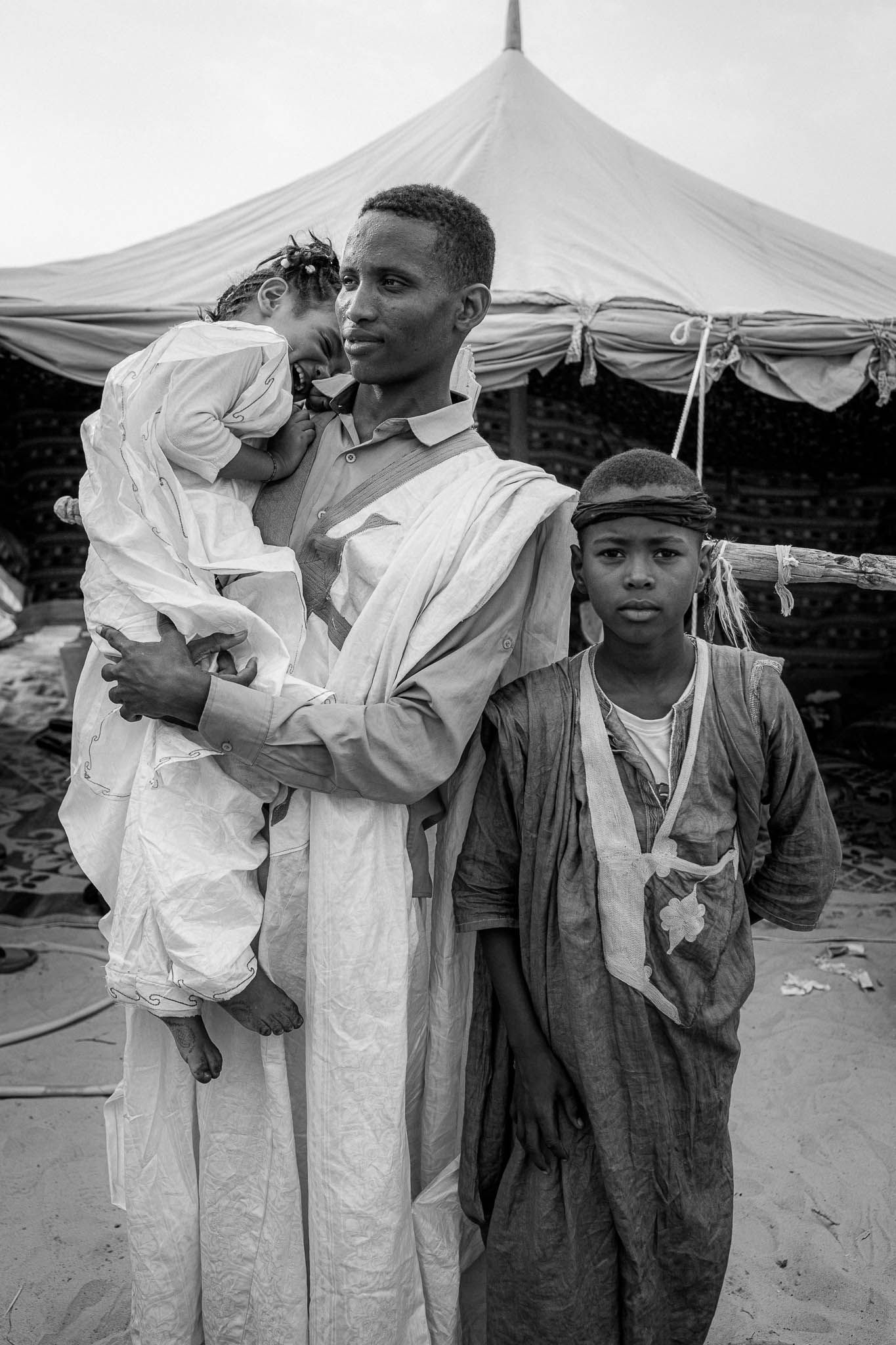 A vintage black and white photograph showing people in traditional African dress standing under a tent canopy.
