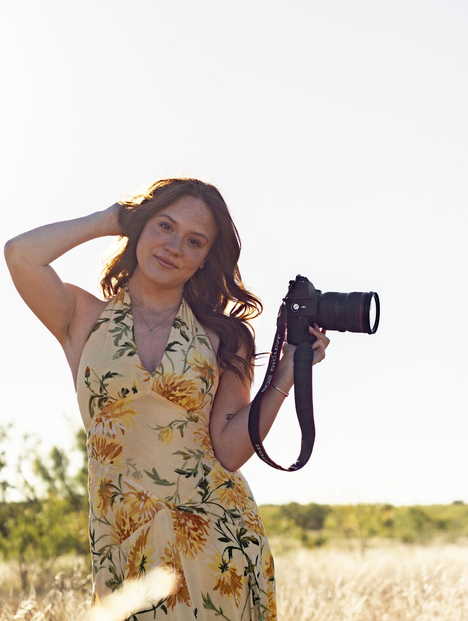 Woman in floral yellow dress posing with DSLR camera in open field under bright clear sky.