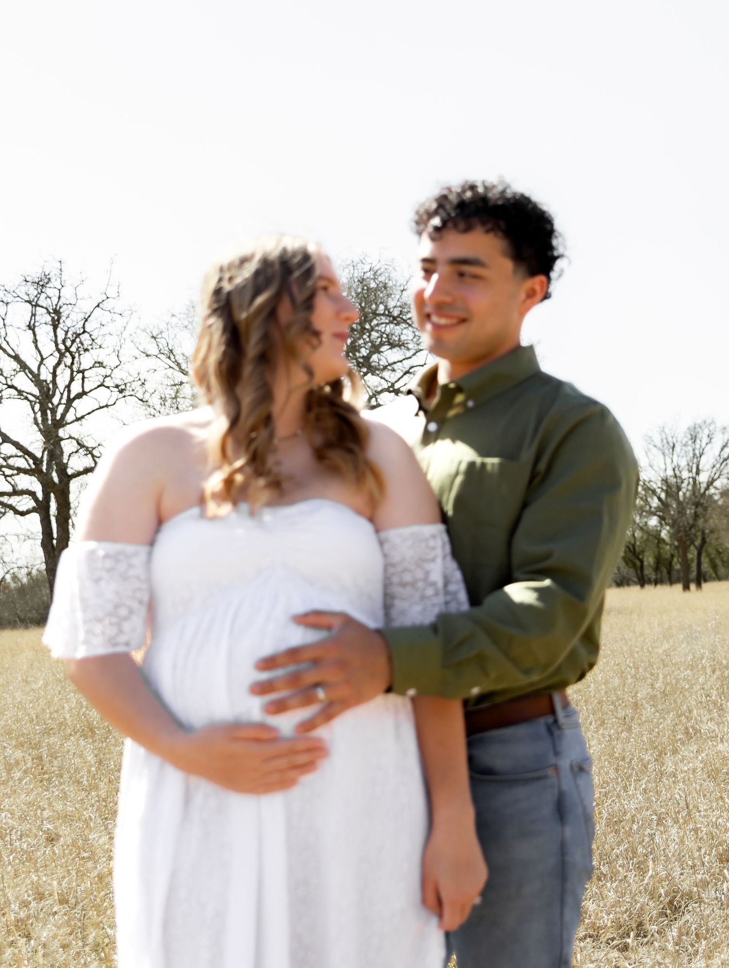 Smiling couple in golden field, man in green shirt embracing pregnant woman in white dress.