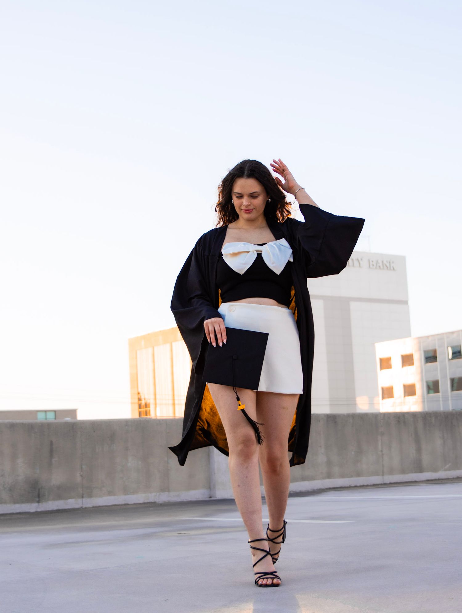 Woman in graduation gown with white bow top and shorts posing on rooftop at dusk.