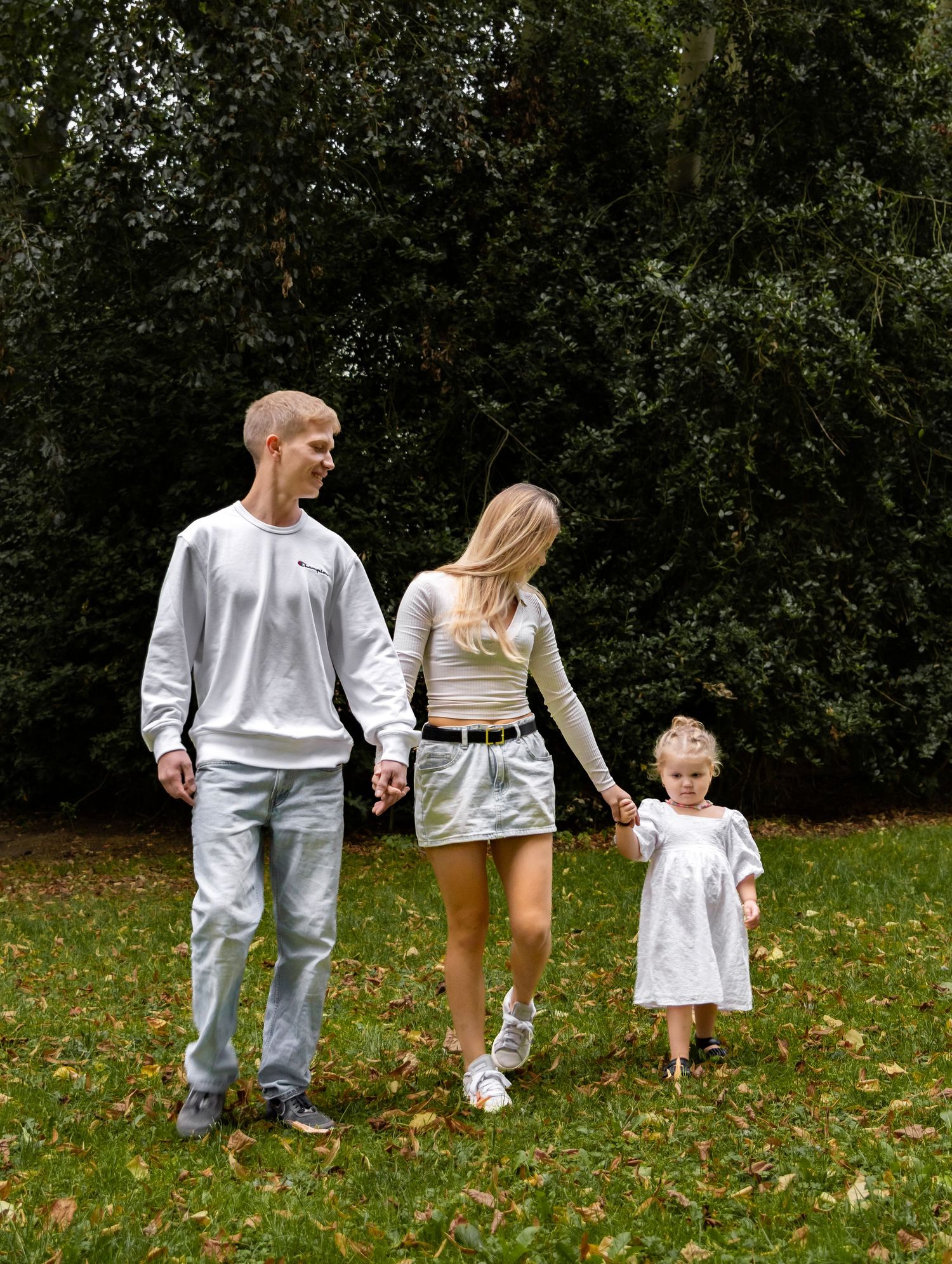 Young family of three walking hand in hand across a green lawn with dense hedges behind them.