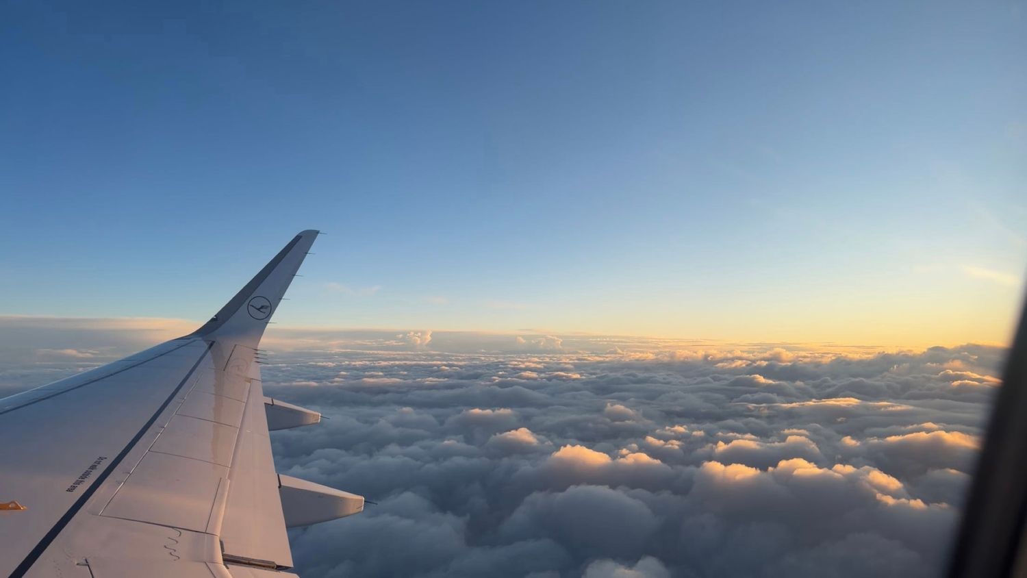 Airplane wing soaring above fluffy white clouds during a warm golden sunset sky.