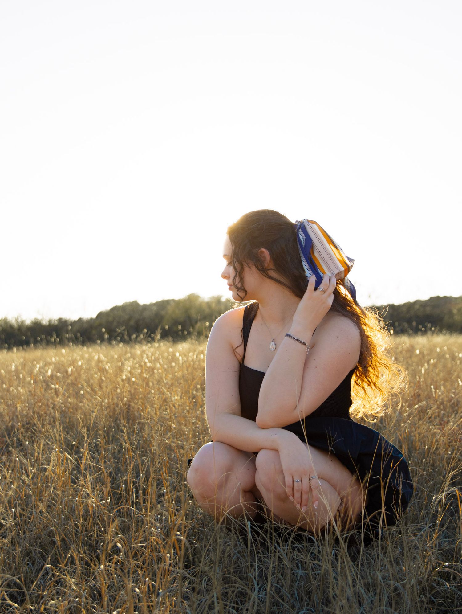 Young woman crouching in golden field at sunset holding colorful hair accessory in black tank top.