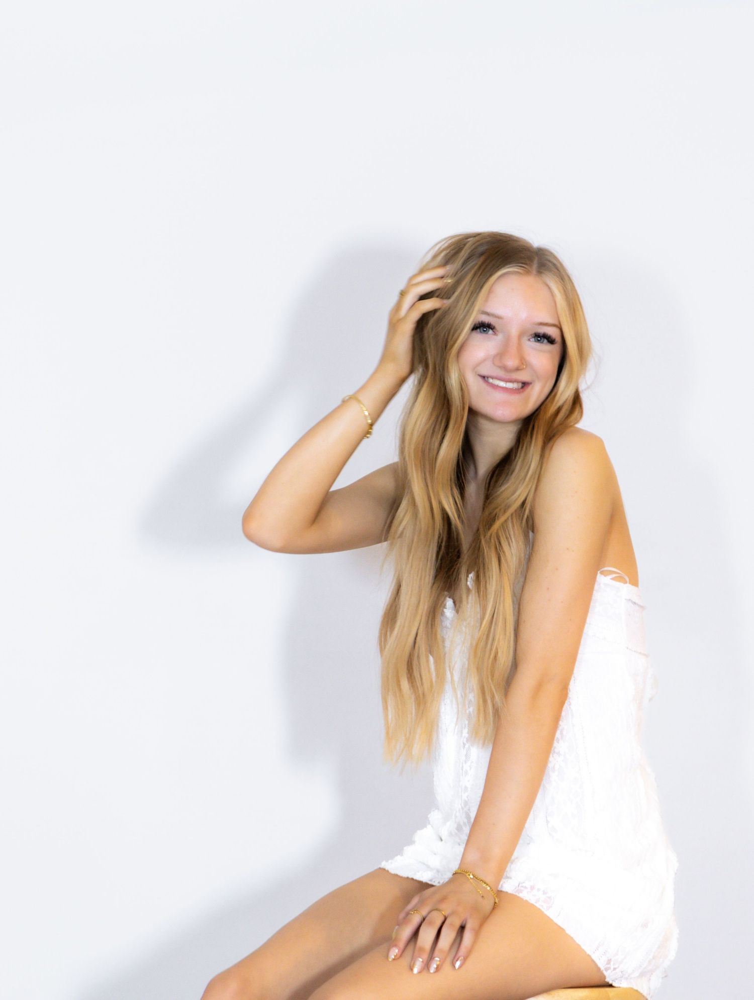 Smiling blonde woman in white romper sitting on yellow stool against white background.