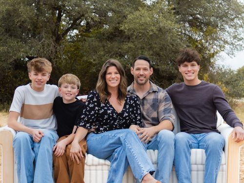 Happy family of five sitting together outdoors with green trees in background on sunny day.