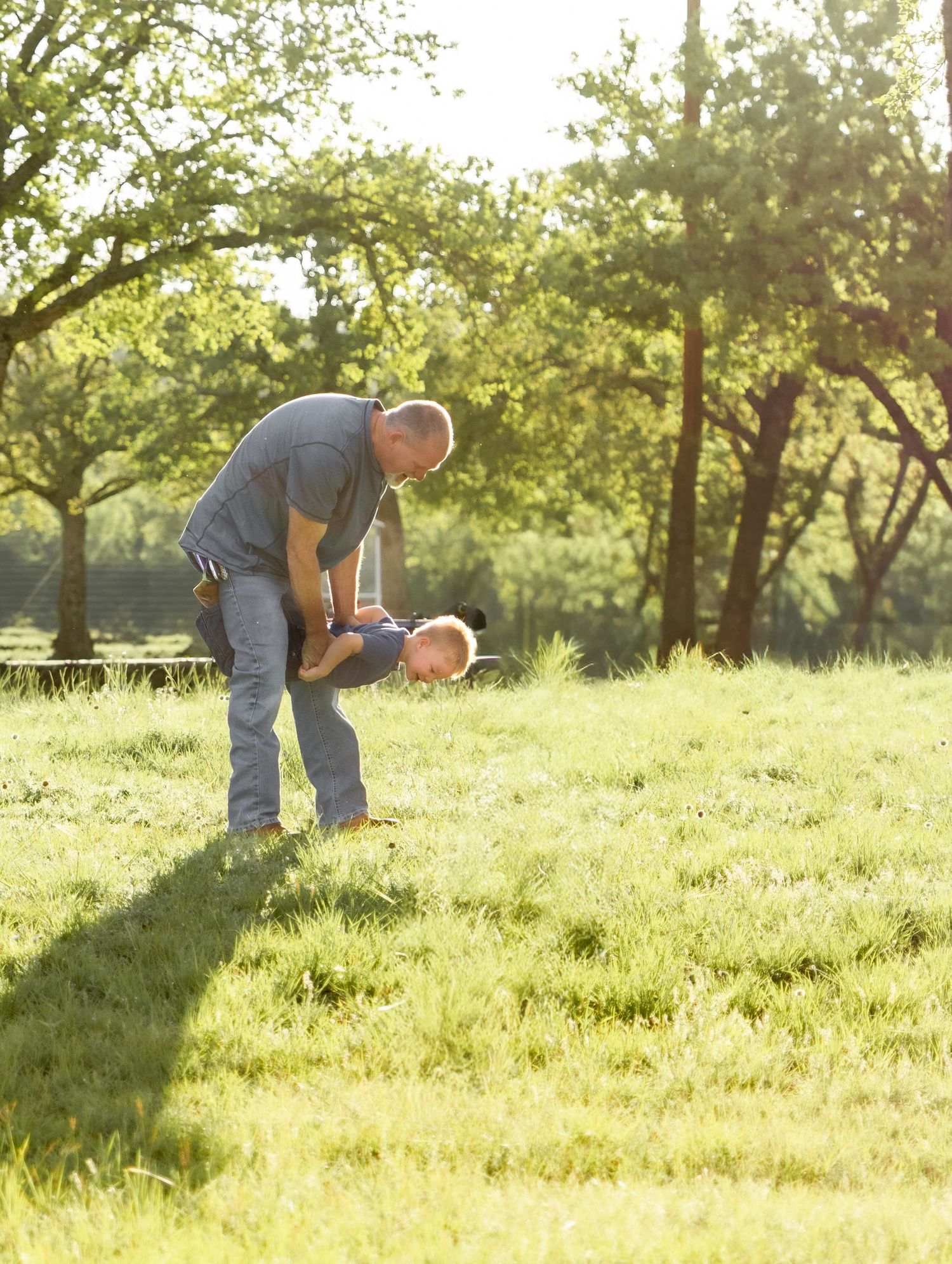 Father playfully holding toddler upside down on a bright sunny green lawn during family photos.
