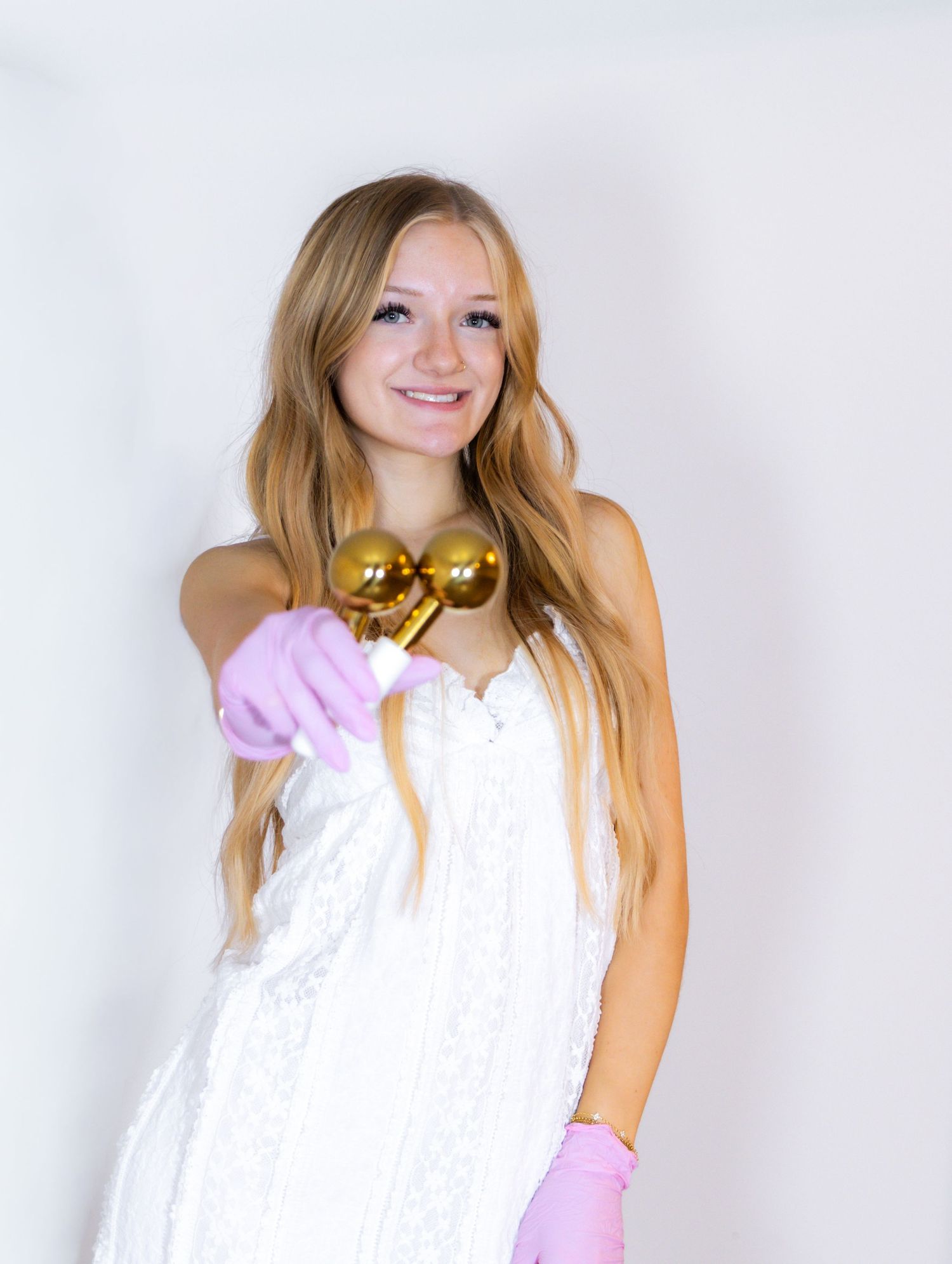Smiling young woman in white lace dress holding gold ornaments wearing pink gloves on white background.