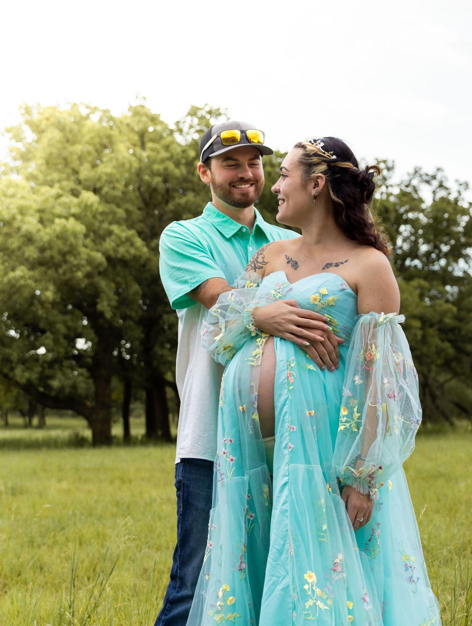 Smiling couple in green field, woman in teal maternity gown with floral embroidery.