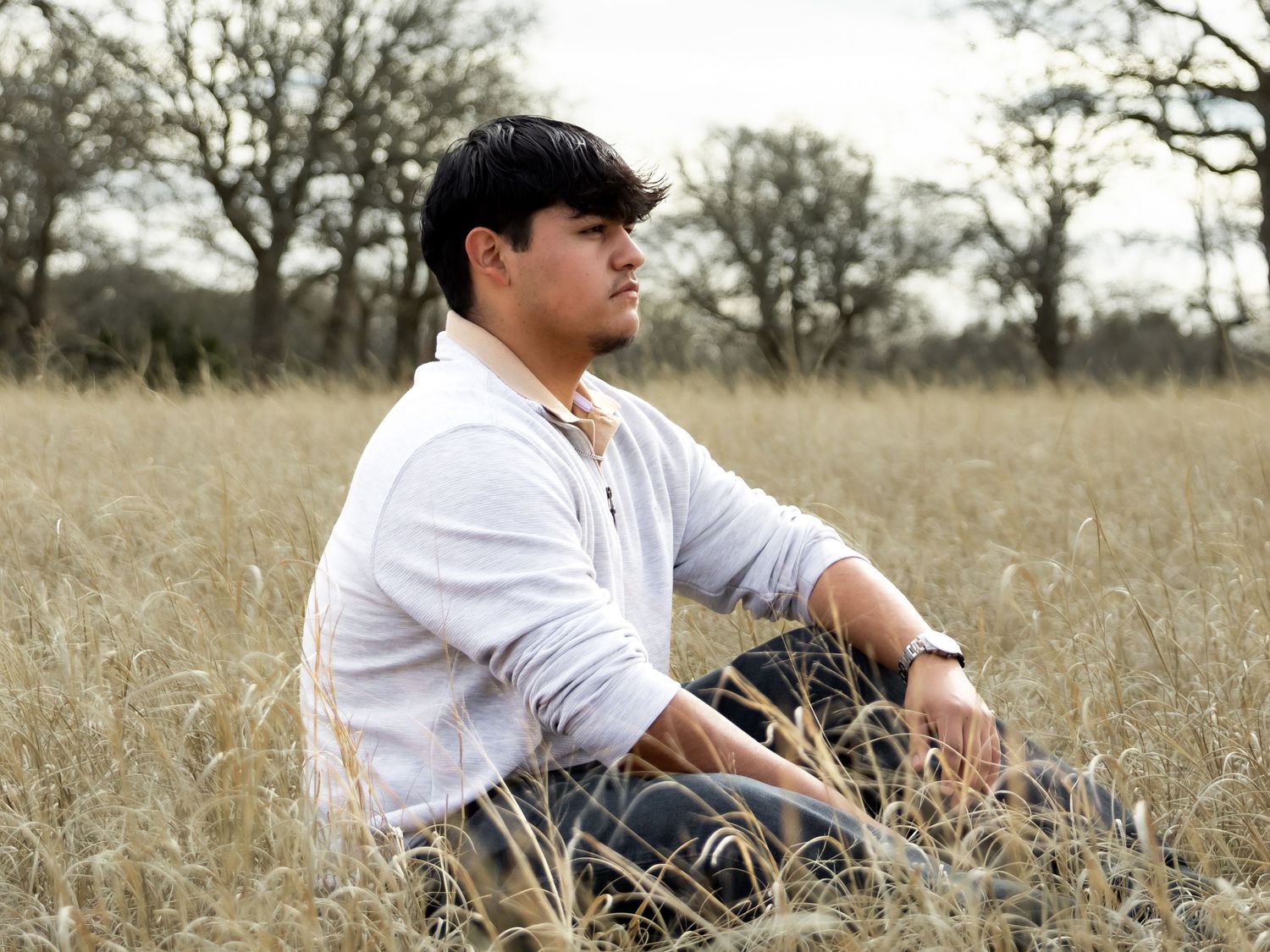 Young man in white hoodie and jeans sitting relaxed in golden grass field with trees.