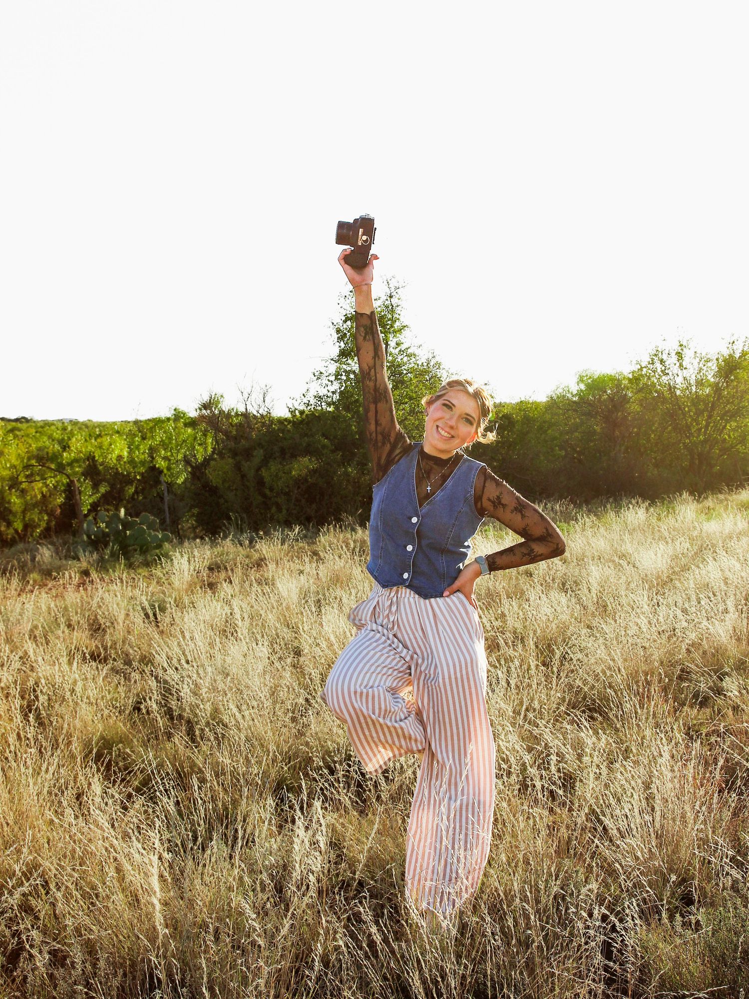 Joyful woman jumping in golden field holding camera up celebrating outdoors at sunset.