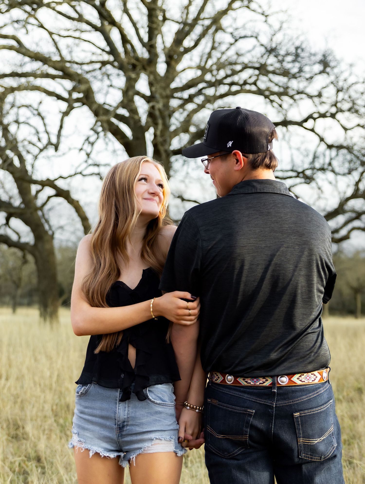 Young couple holding hands in a grassy field with bare trees, woman smiling at man in black cap.