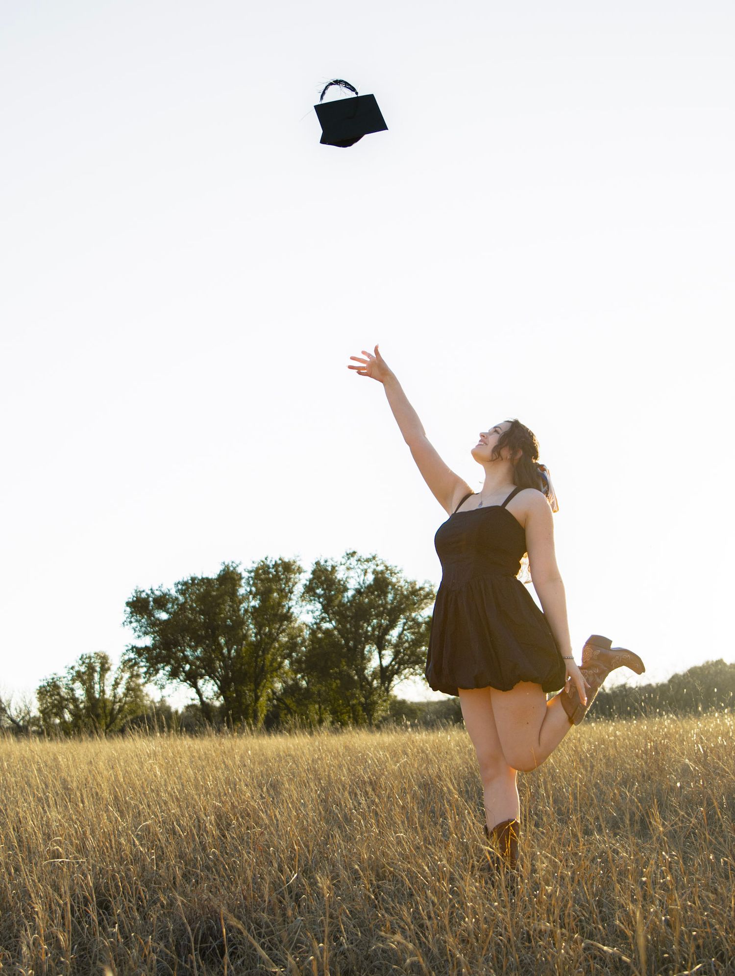 Joyful woman in black dress tossing graduation cap in golden field wearing cowboy boots.