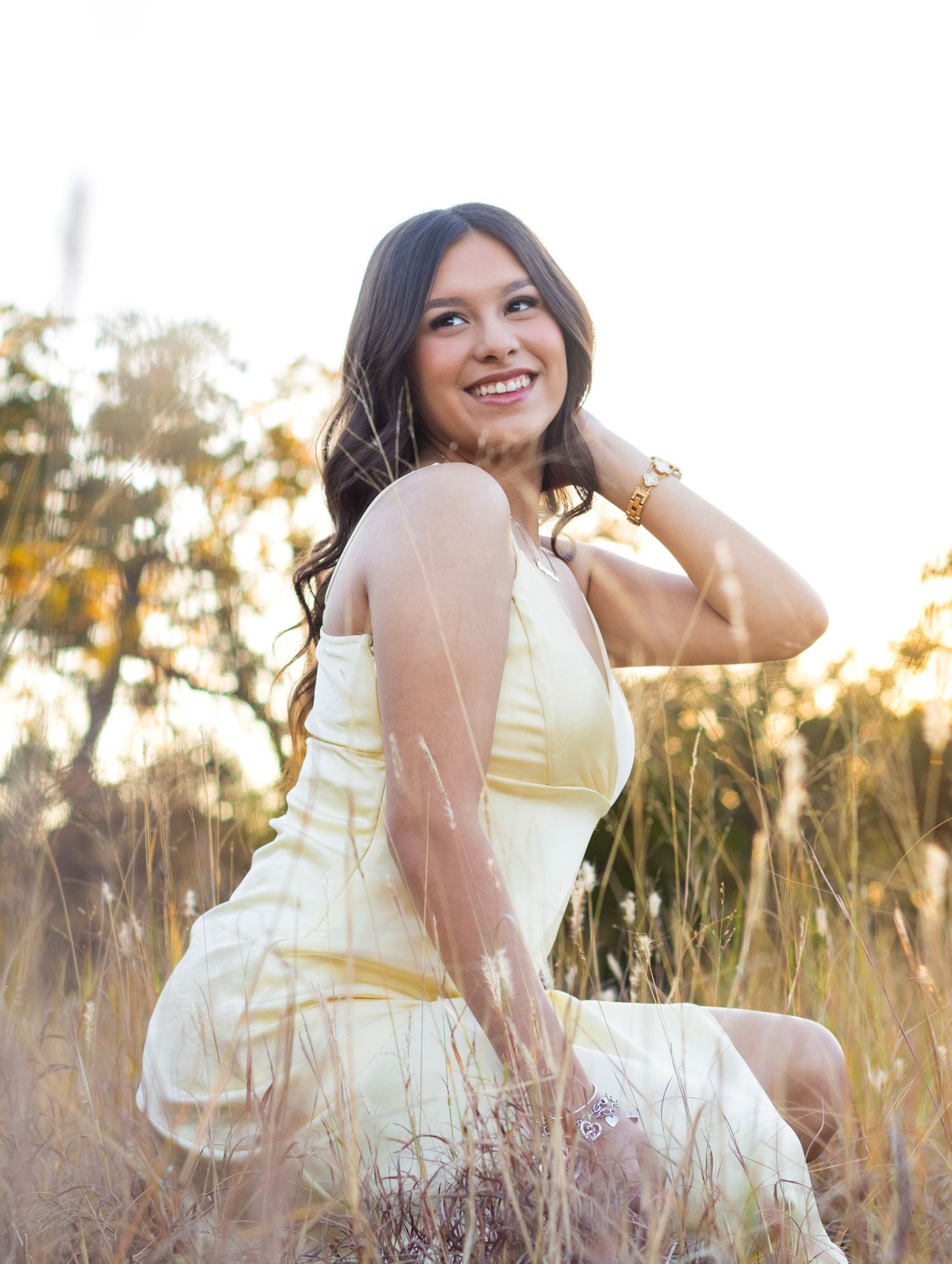 Smiling young woman in yellow dress sitting in sunlit golden field touching her hair.