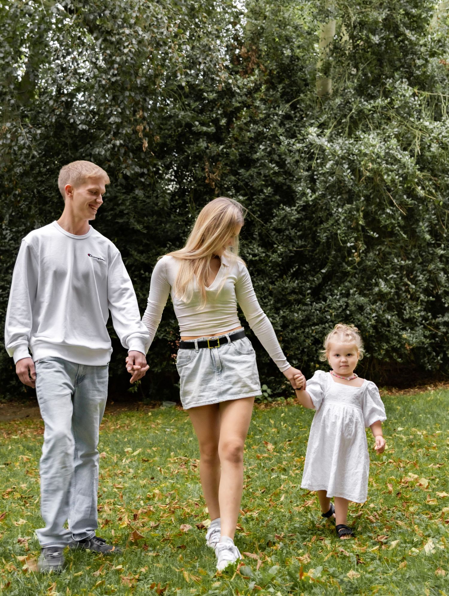Happy young family holding hands walking together on grass surrounded by green trees.