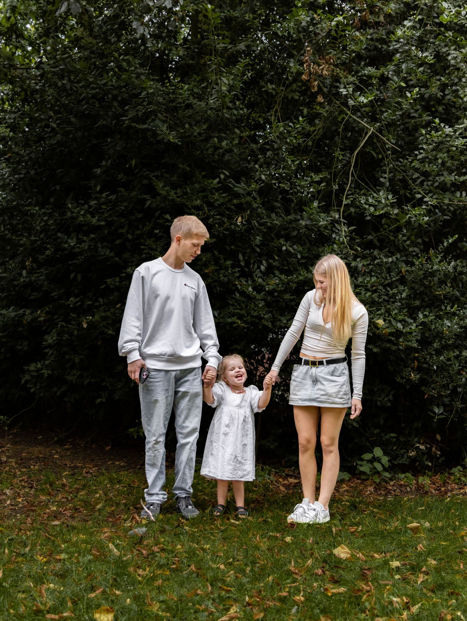 Young family with toddler girl walking hand in hand on green lawn in front of lush bushes.