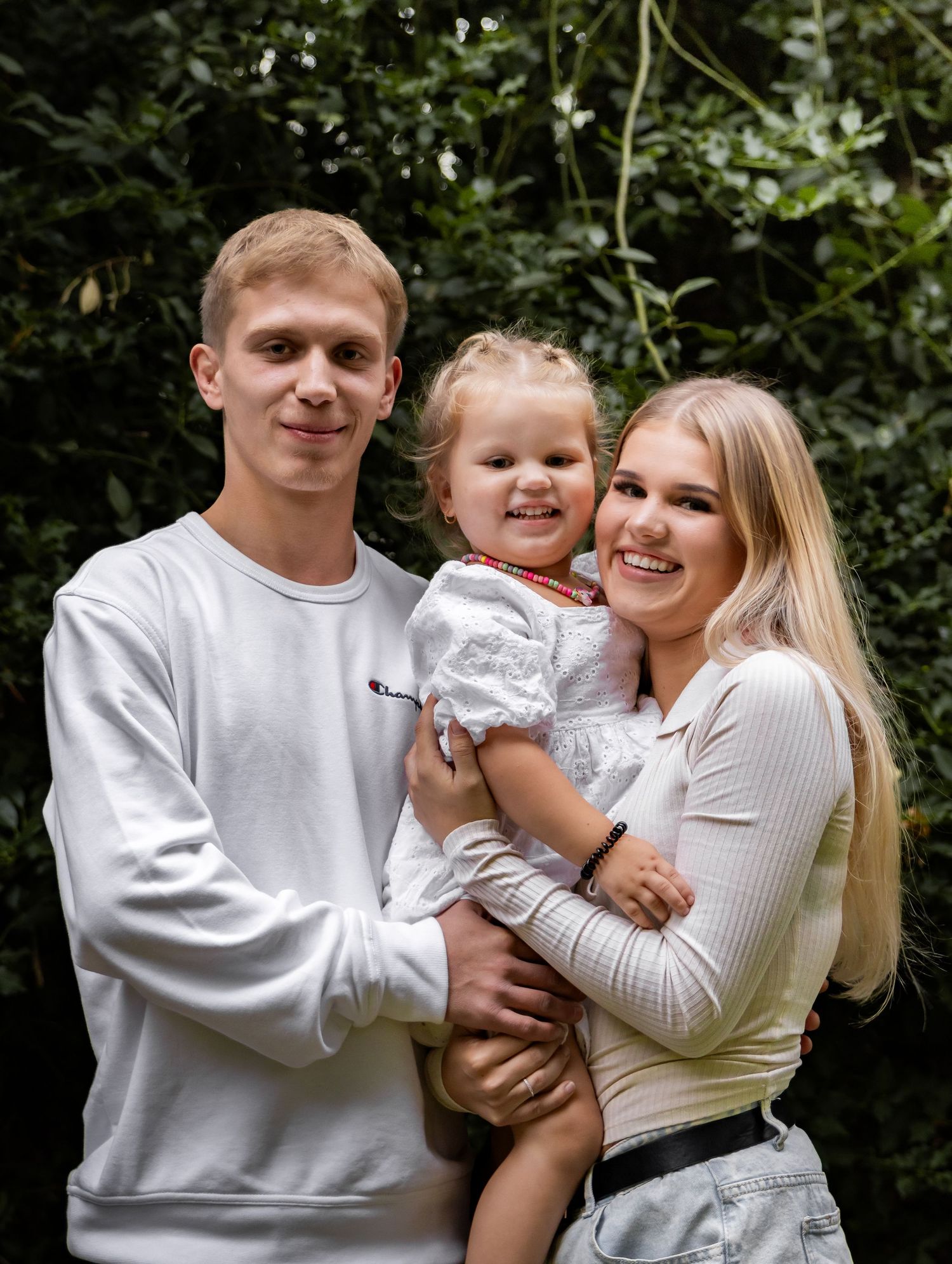 Smiling young family with blonde toddler posing together outdoors against green leafy background.